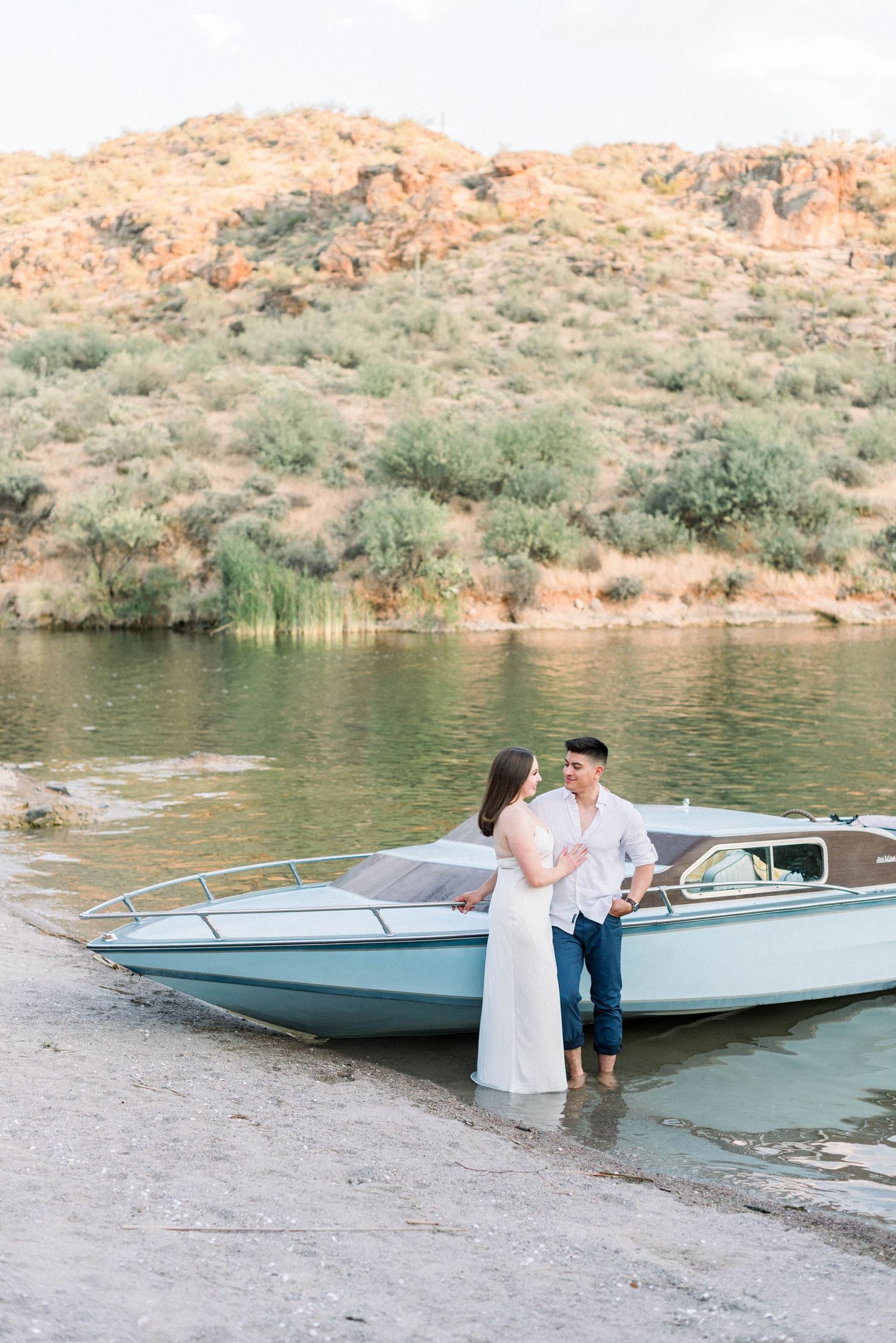 Couple in white sharing a romantic moment by a boat at a desert lake.