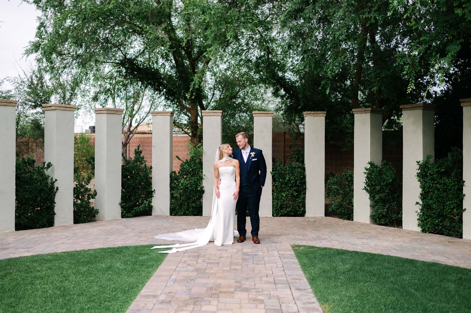 Bride and groom stand together in an elegant outdoor garden courtyard with white columns and lush greenery.