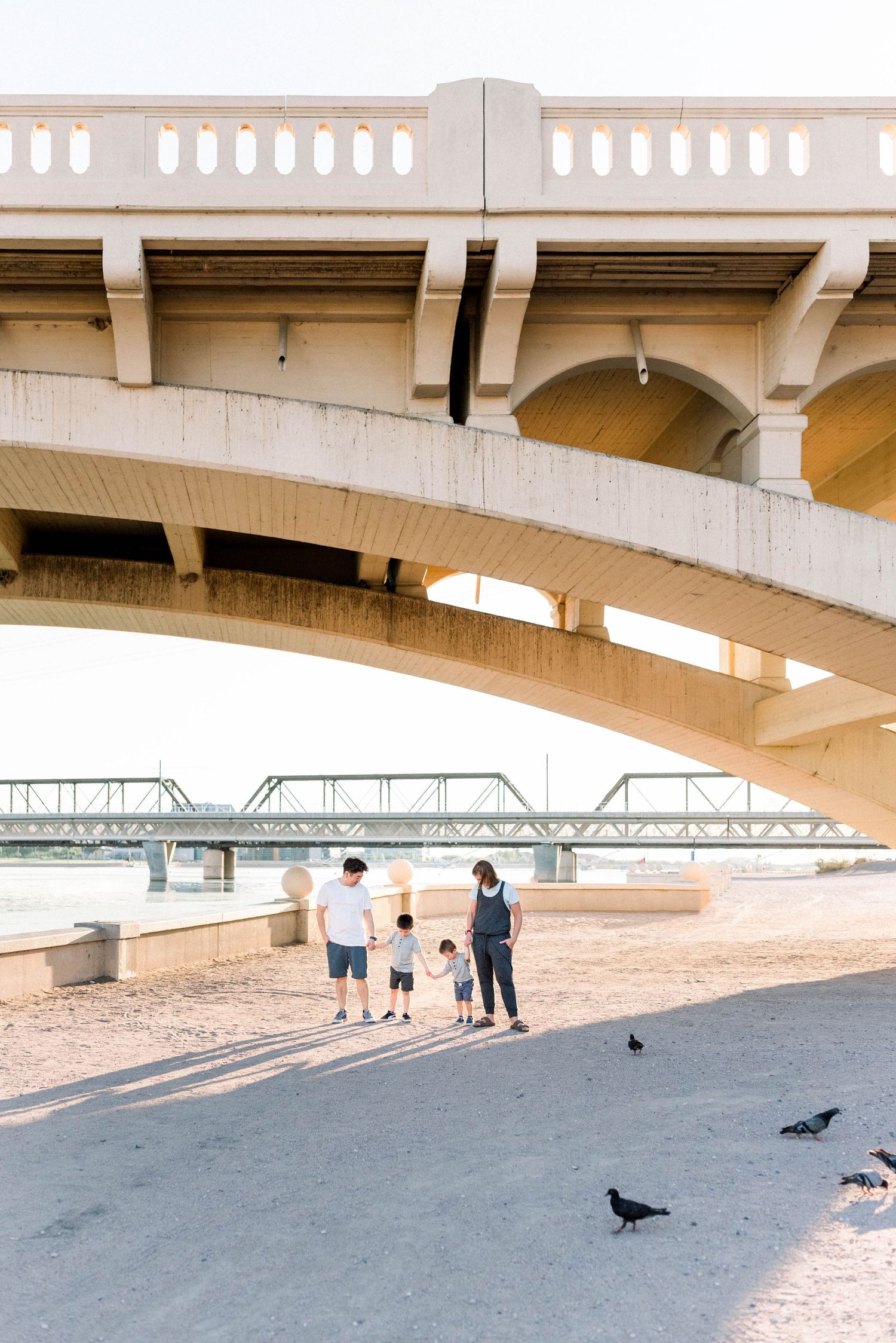 A group stands beneath a concrete bridge overpass while pigeons gather on the pavement nearby on a sunny day.