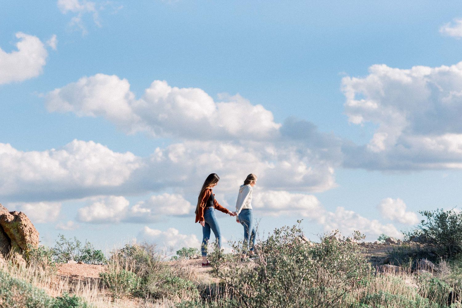 People walking along a scenic hilltop trail with white puffy clouds in a bright blue sky above.