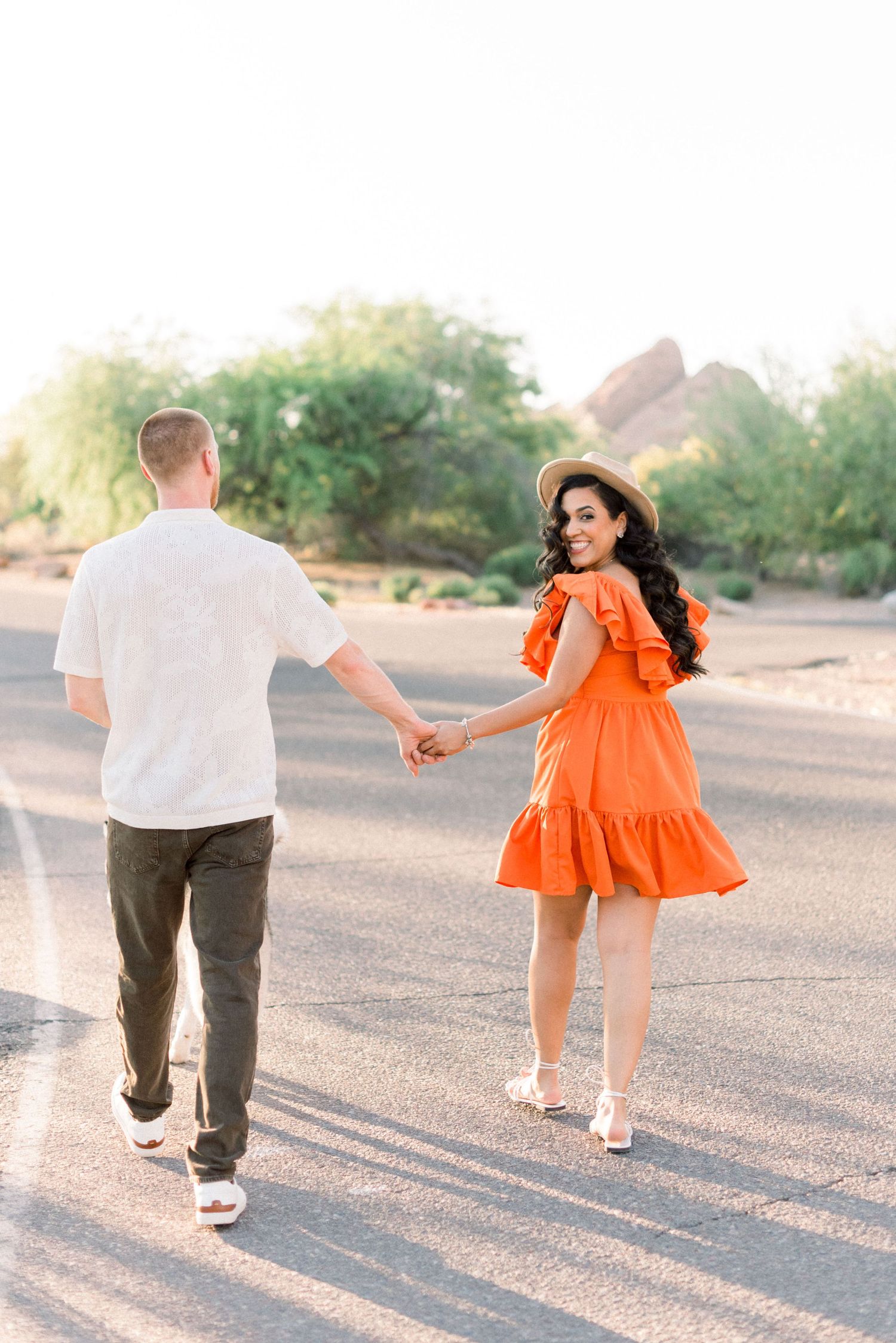 A couple in orange and white outfits dances playfully together in a desert setting during sunset.