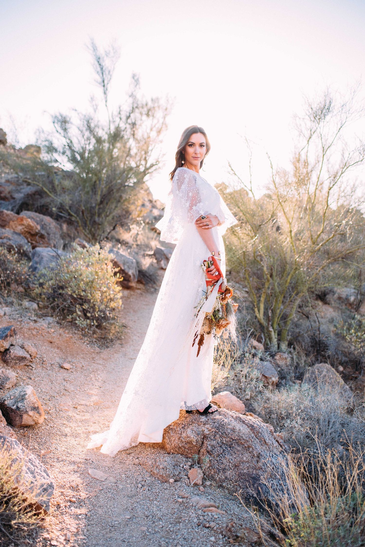 A bride in a flowing white dress stands on a desert trail at sunset holding a floral bouquet among desert brush and rocks.