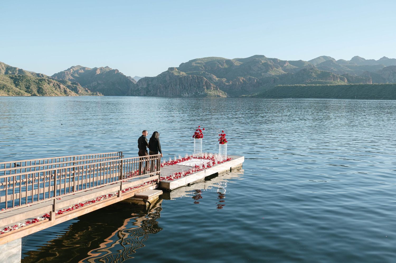 Floating red and white dock extends into calm blue waters surrounded by rugged mountain peaks on a sunny day.