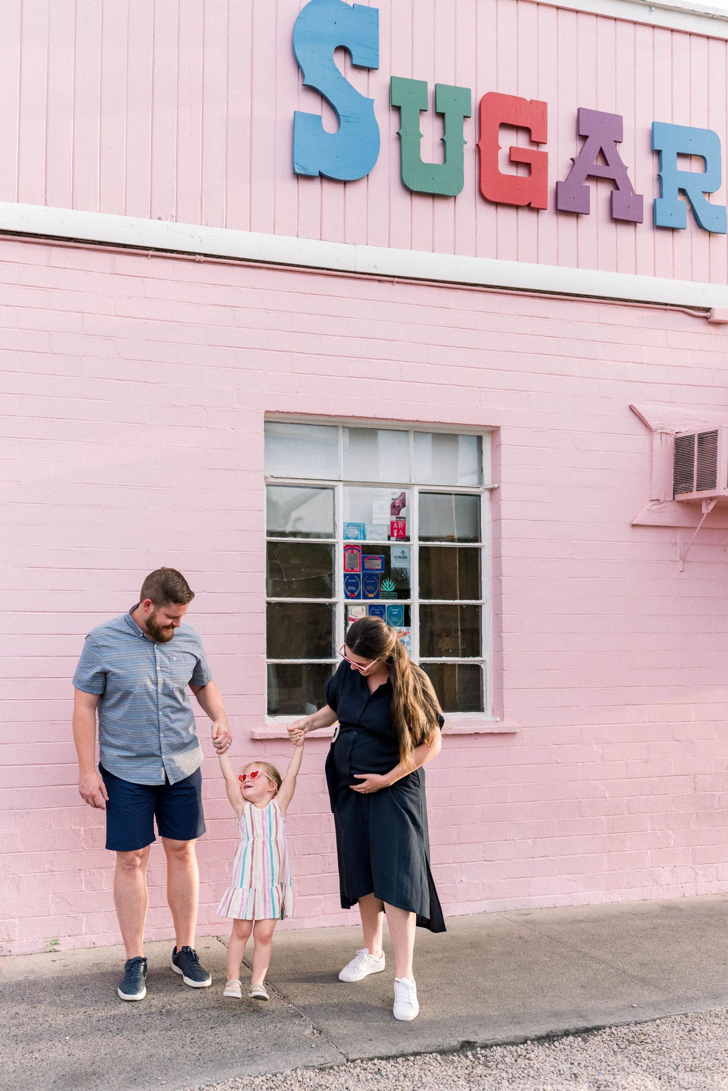 Three people stand outside a pink building with retro blue and red 'SUGAR' signage on the exterior wall.