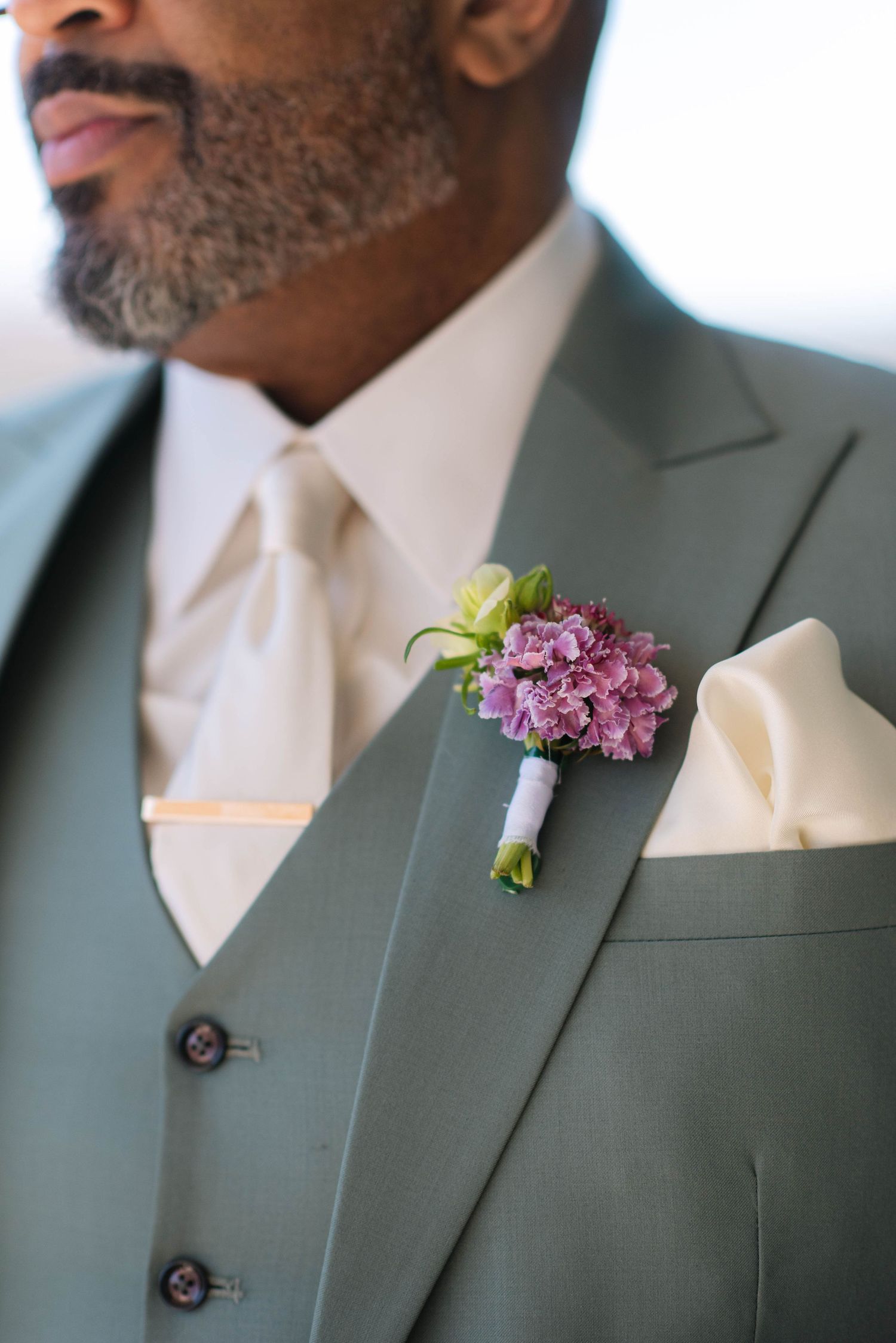 Sage green suit jacket with cream tie and purple floral boutonniere.