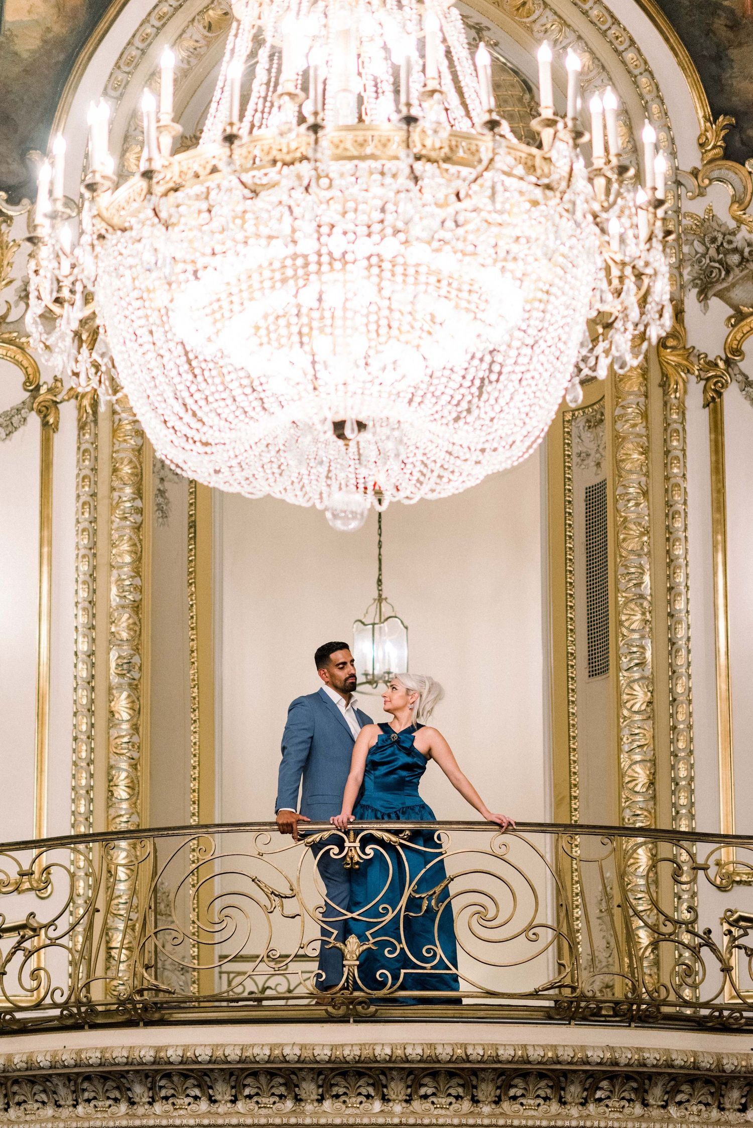 Couple poses dramatically under grand crystal chandelier on ornate balcony.