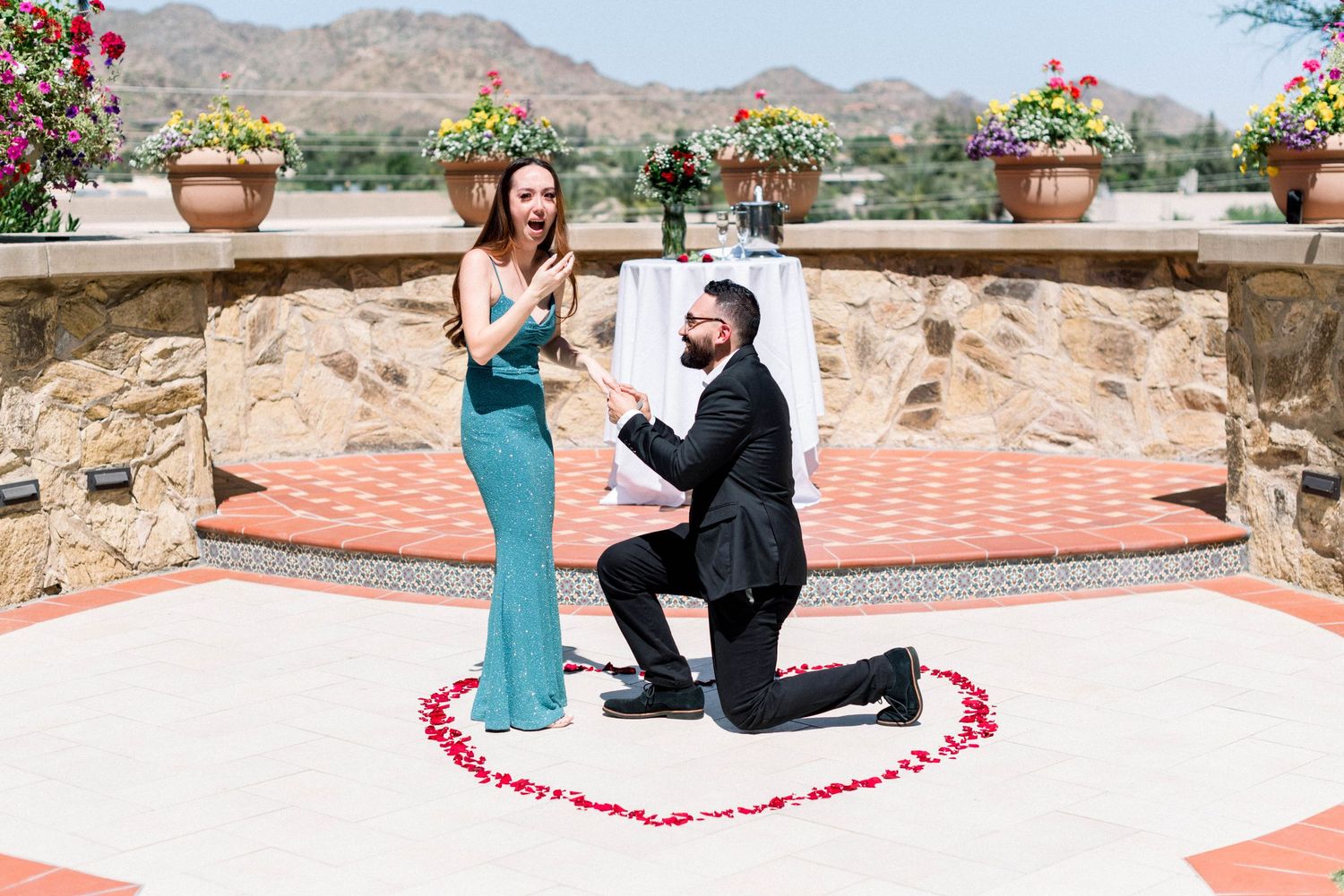 A romantic proposal scene with rose petals arranged in a heart shape on a stone patio with mountain views.