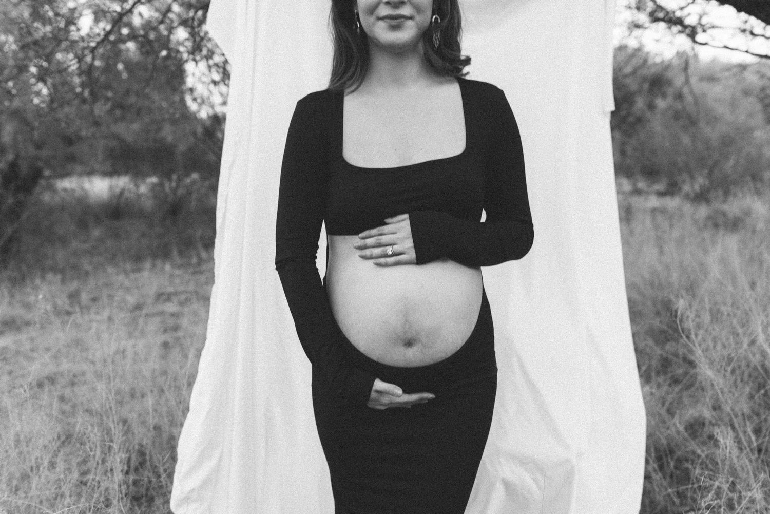 Black and white maternity portrait with natural lighting against white backdrop in field setting.