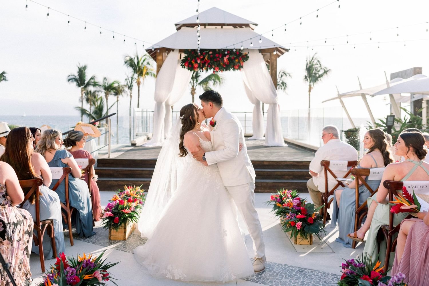 A beautiful outdoor wedding ceremony with guests seated around a decorated gazebo by the beach.