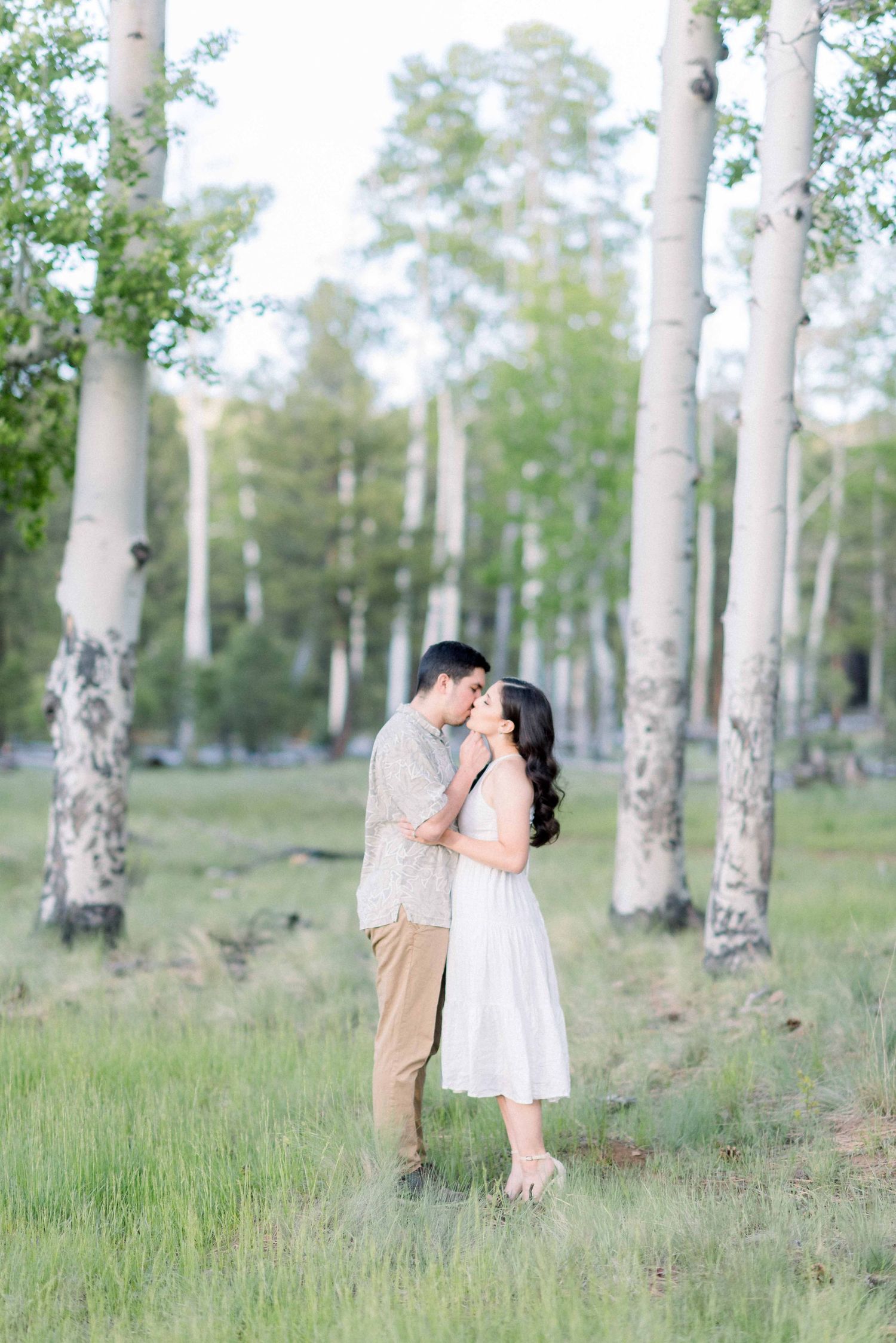 A couple shares an intimate moment among birch trees in a sunlit forest meadow.