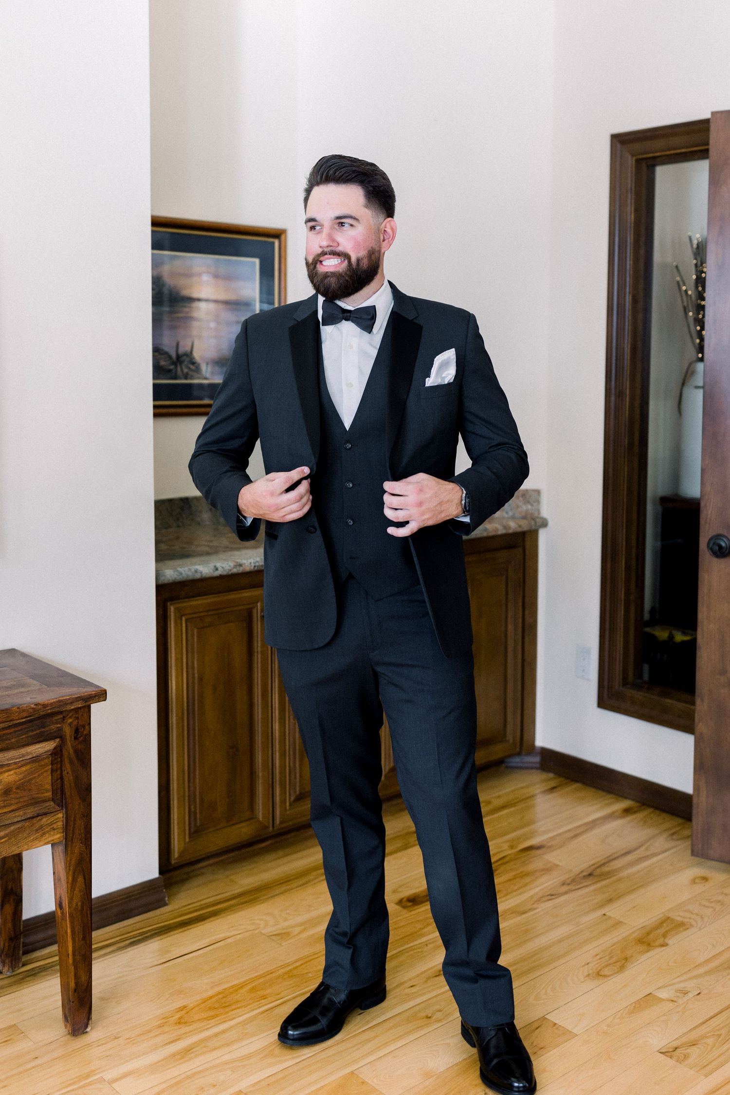 Groom in classic black tuxedo and bowtie poses confidently in formal setting.