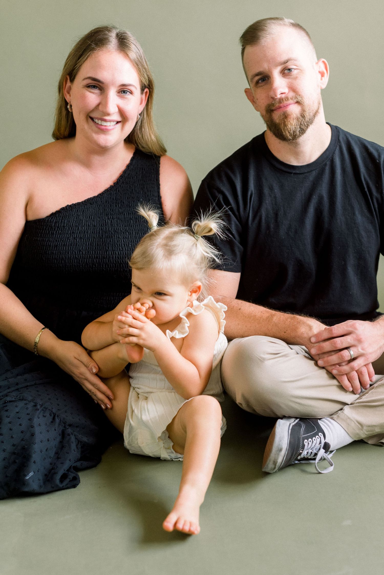 Family portrait with parents and toddler sitting together against neutral backdrop.