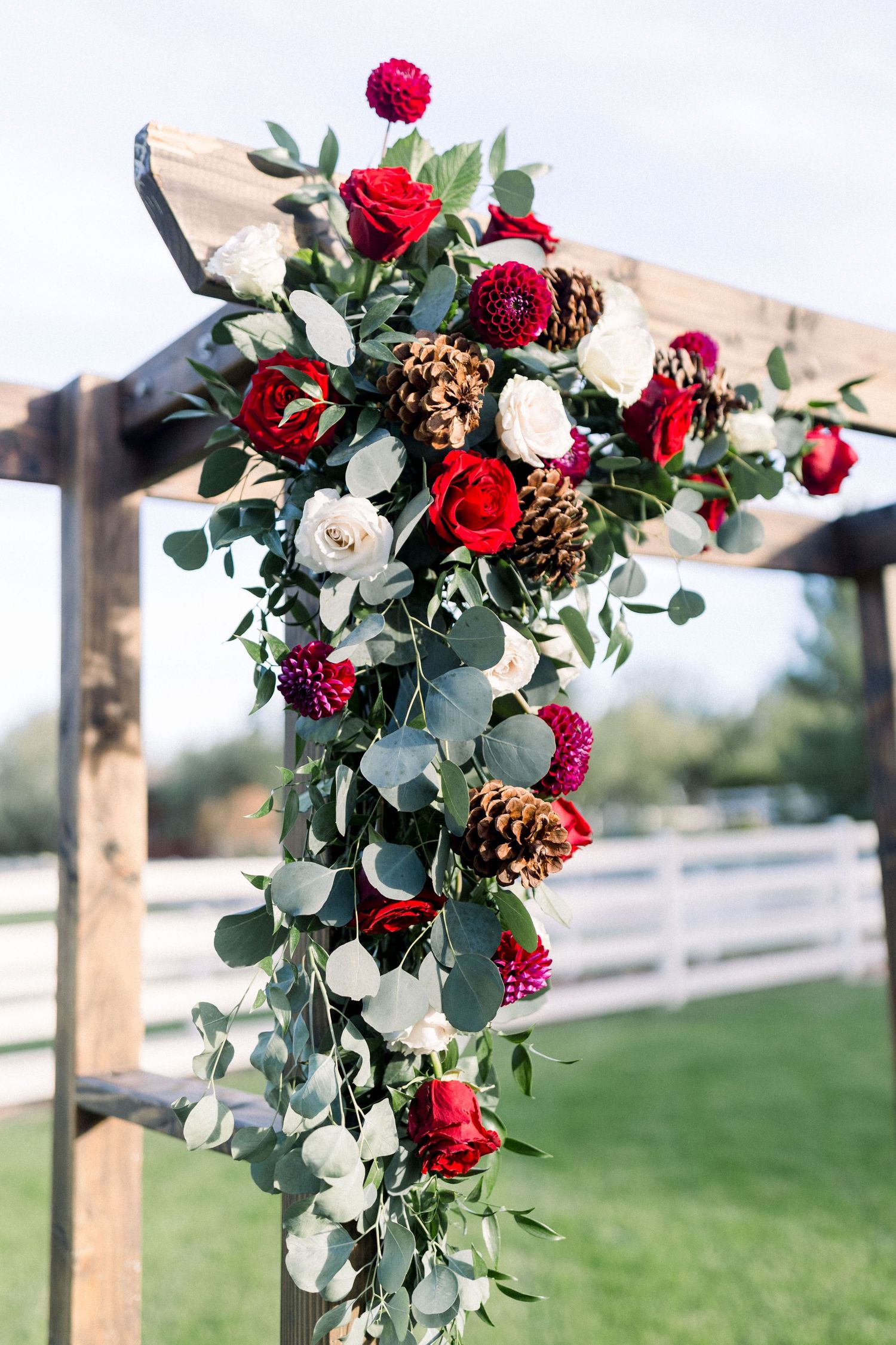 Wedding arch detail showing cascading red roses, pine cones, and eucalyptus greenery.