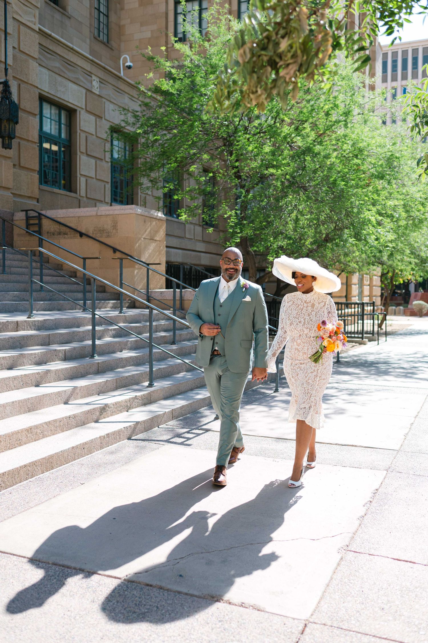 A couple walking down stone steps outside a historic building on a sunny day, casting long shadows on the pavement.