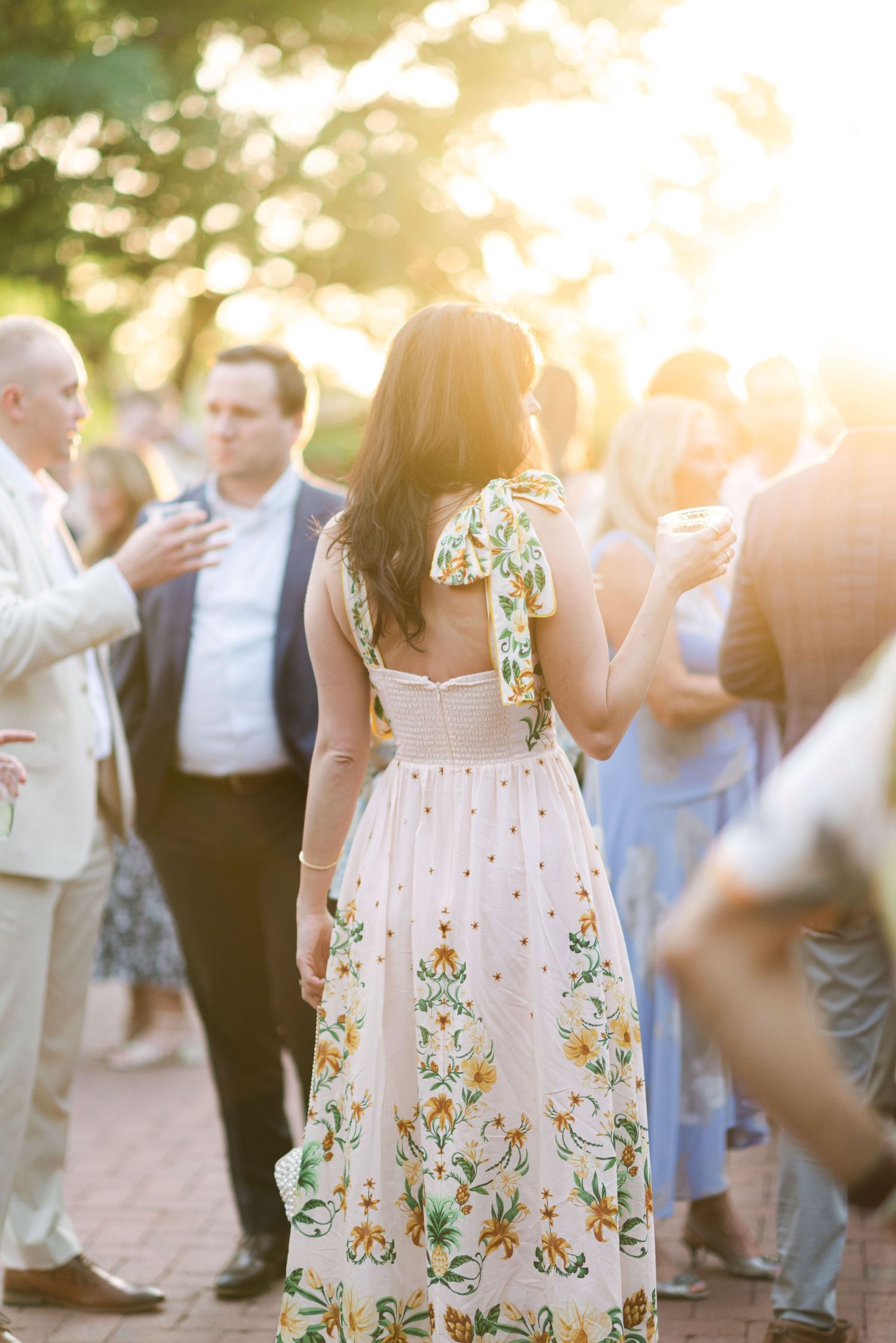 Wedding guests gather outdoors at sunset with someone wearing a floral summer dress in focus.