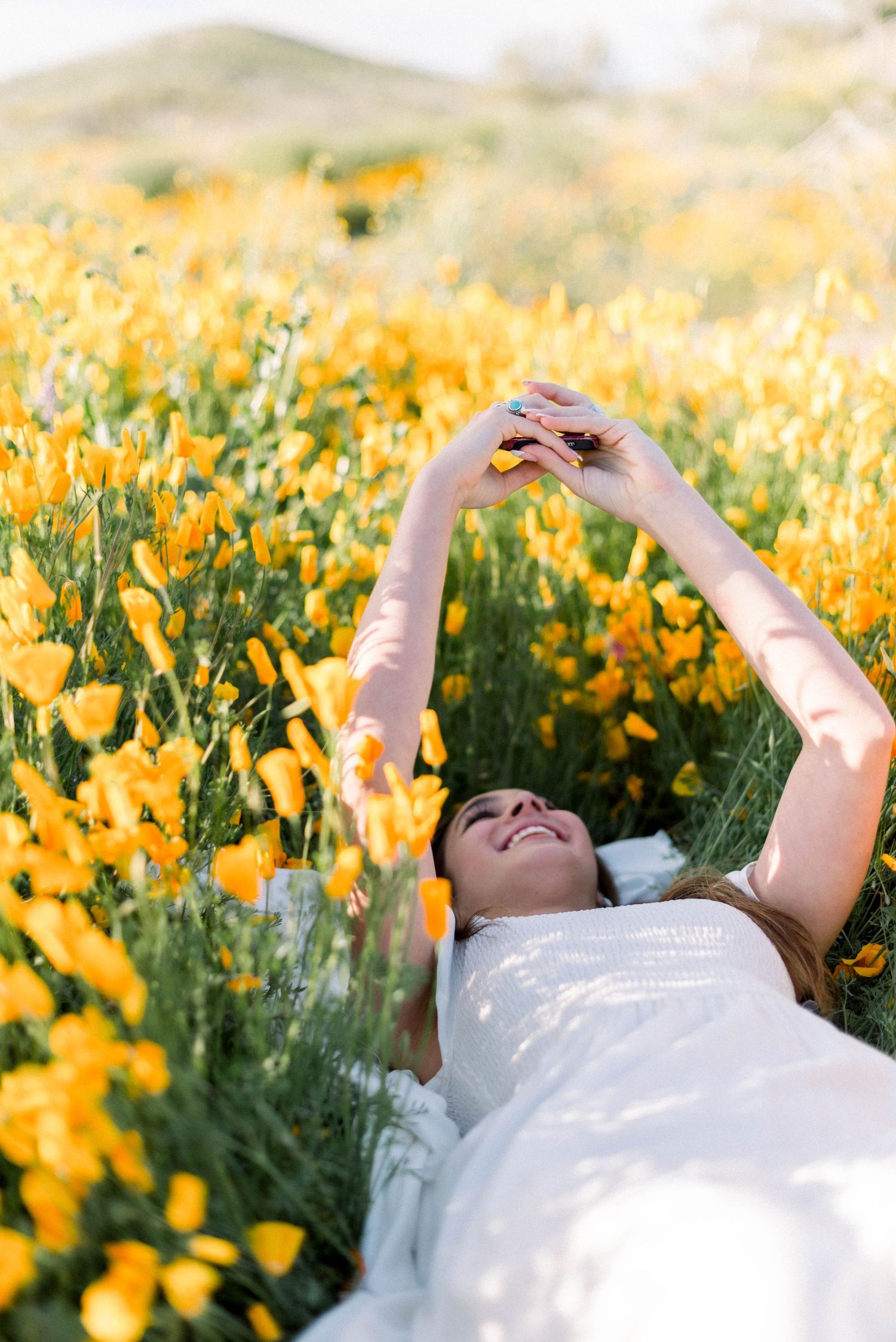 Laying in golden wildflower field wearing white dress with arms raised joyfully.