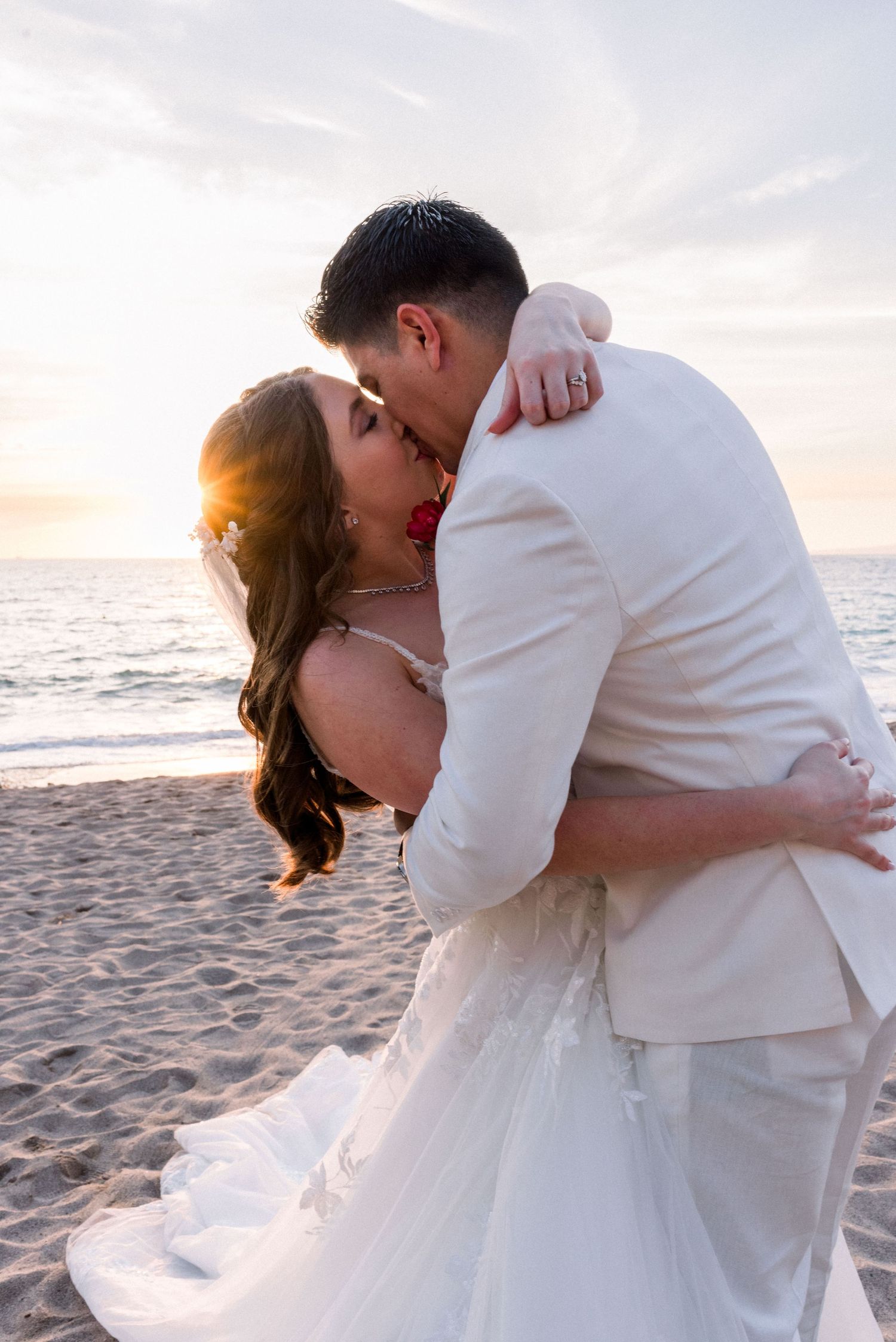 Newlyweds share an intimate moment on the beach at sunset with ocean waves in the background.