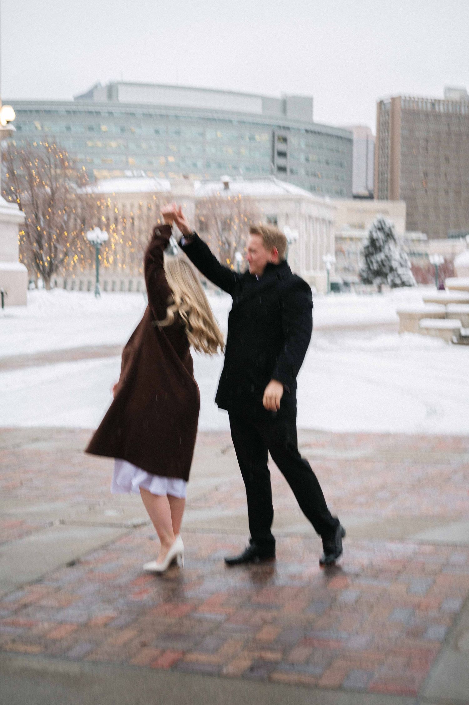 A couple dances playfully in the snow on a brick pathway.