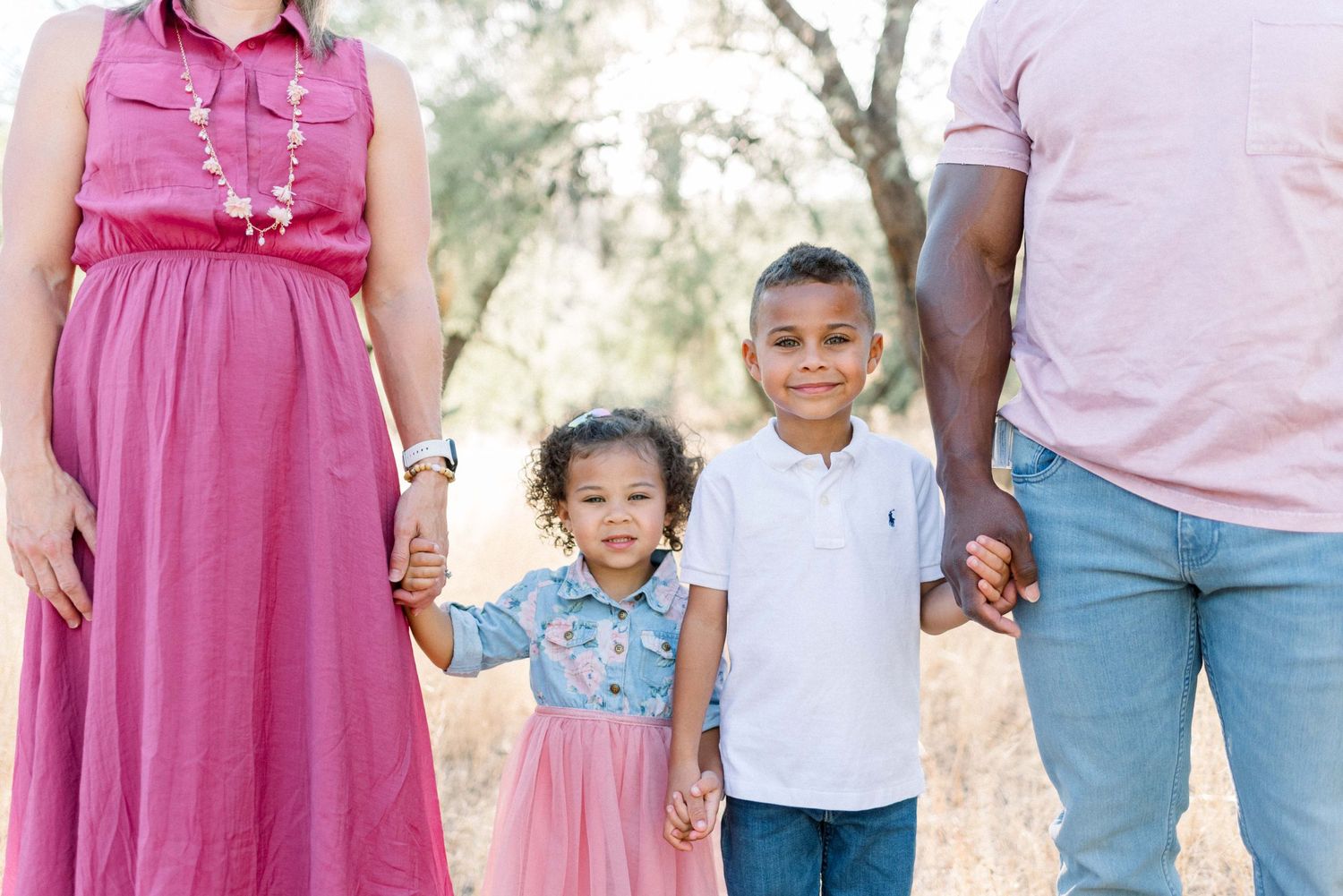 A family holding hands outdoors, with a pink dress and casual attire.