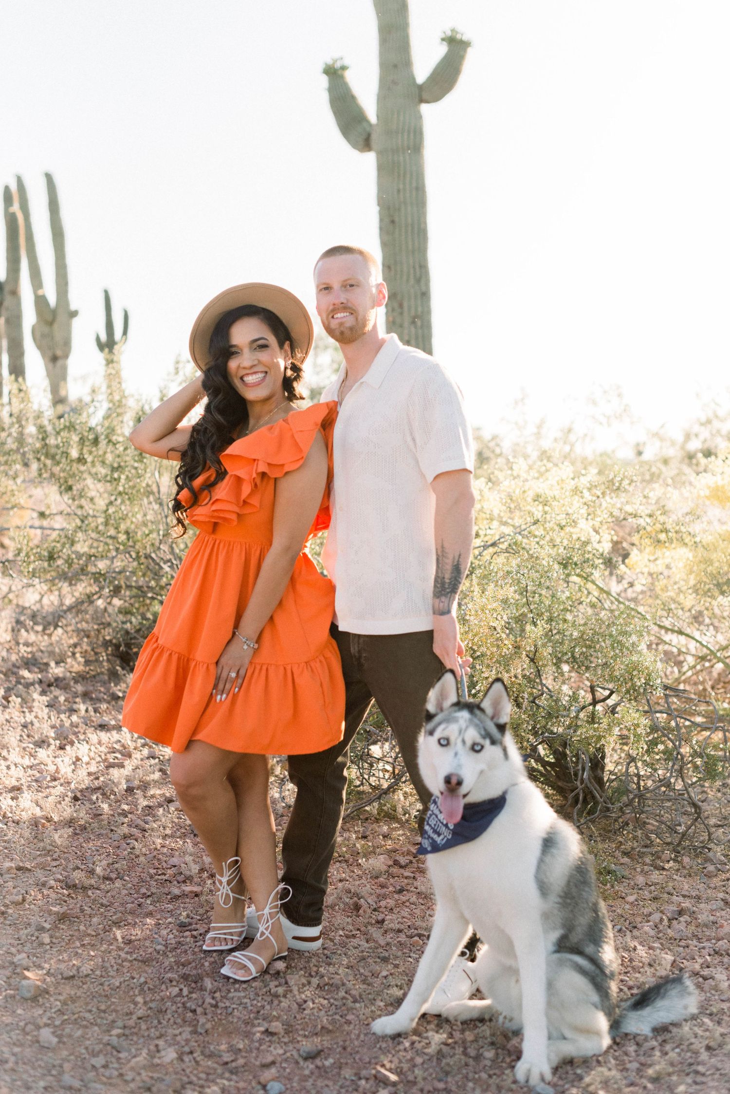 Couple in desert setting with orange dress and husky dog among saguaro cacti.