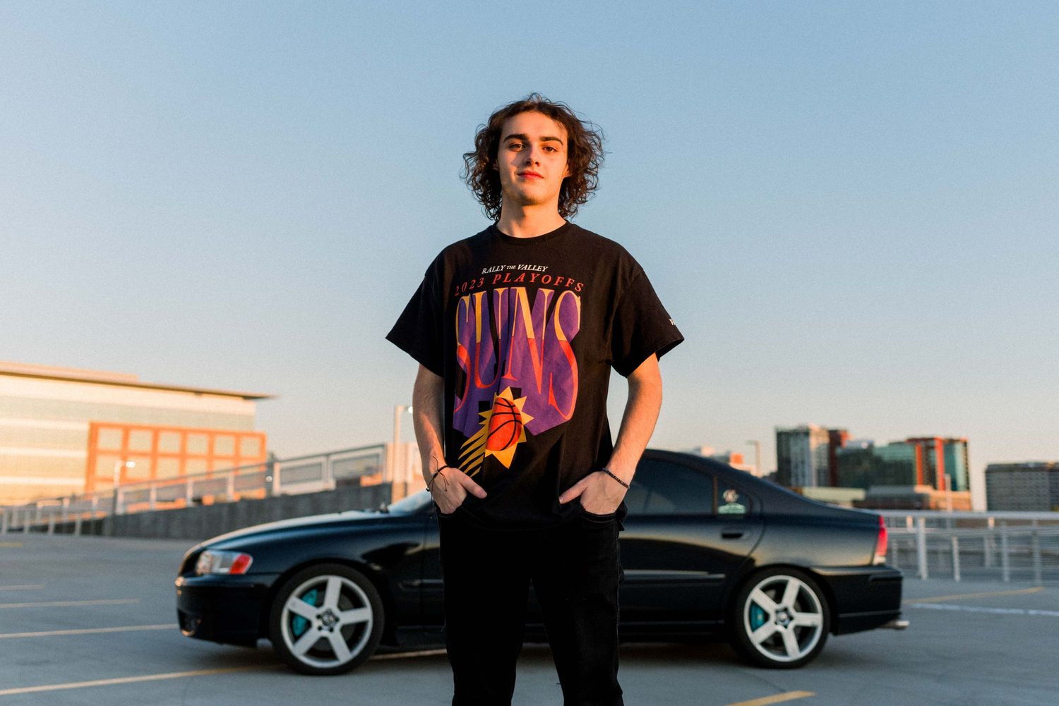 Person in black t-shirt poses with modified black sedan on urban rooftop.