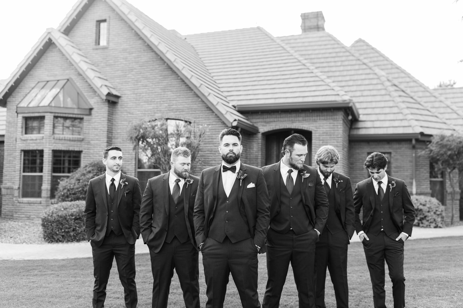 Groomsmen stand in a line wearing dark suits in front of a brick house in black and white.