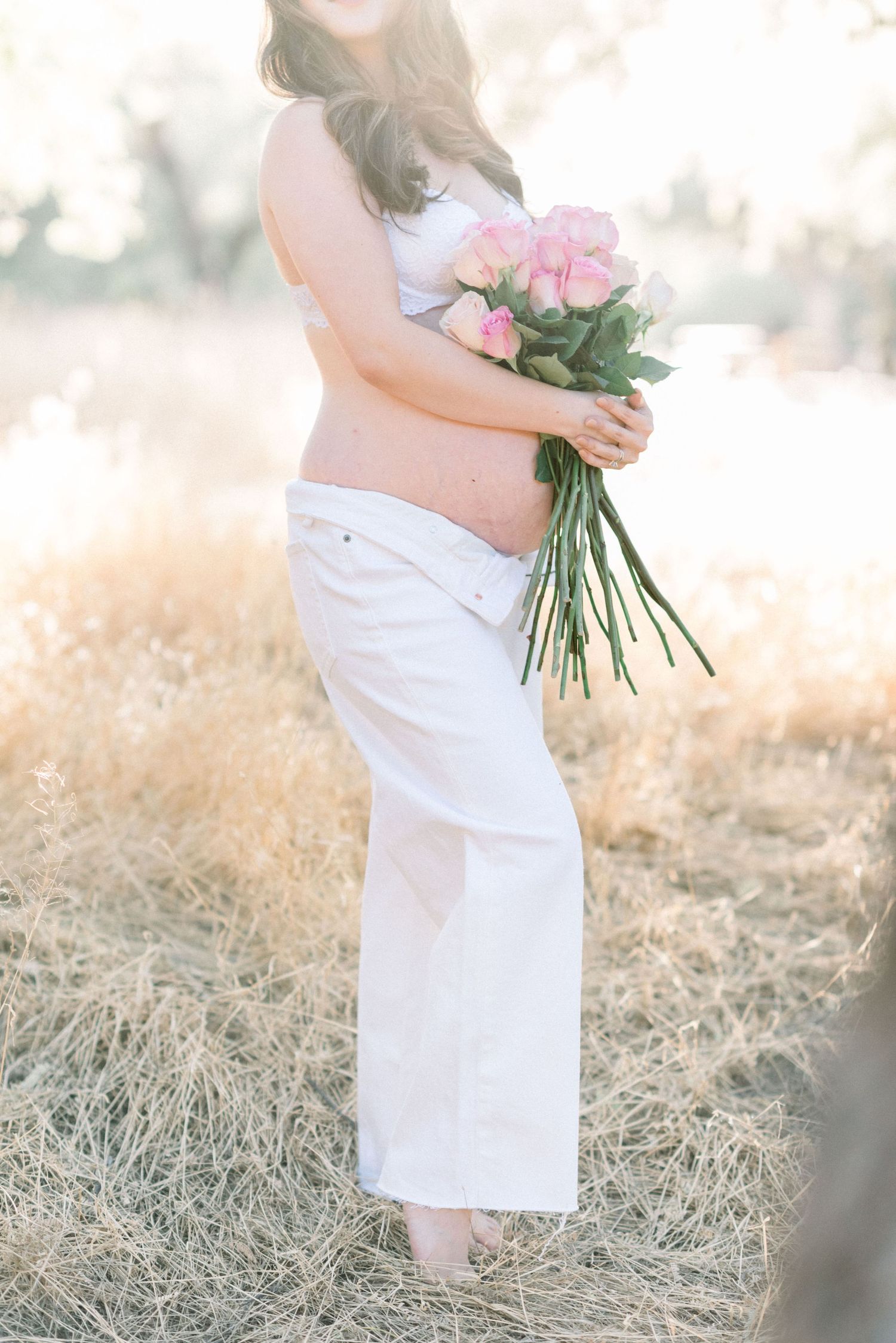 Expectant mother in white dress holds pink roses while standing in golden field during sunset maternity photoshoot.