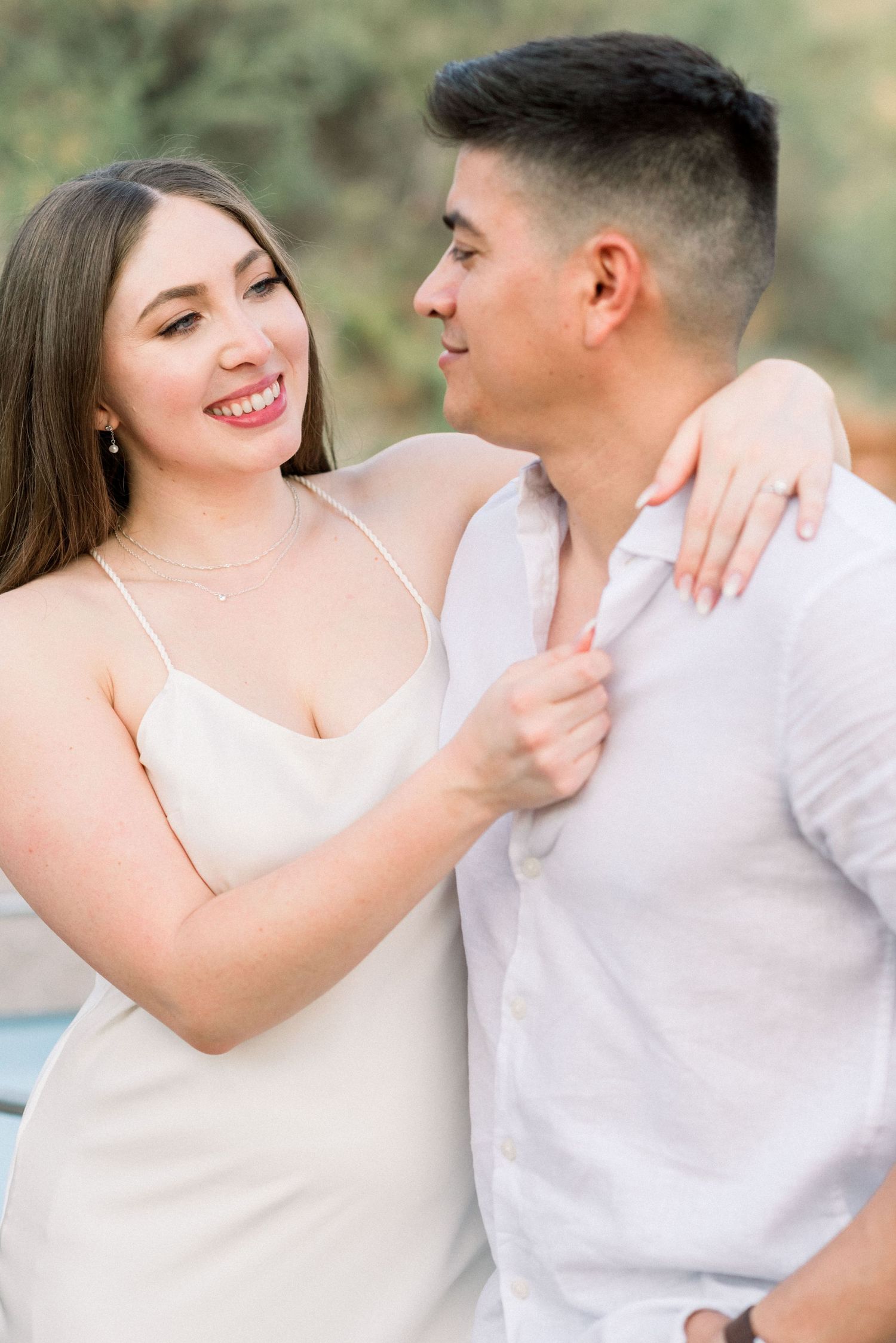 A romantic engagement photo of a couple embracing outdoors in soft natural lighting.