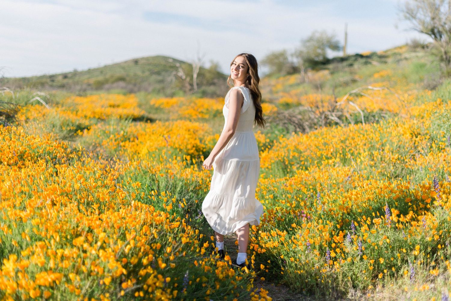 Figure in white dress stands amid rolling hills covered in golden wildflowers.