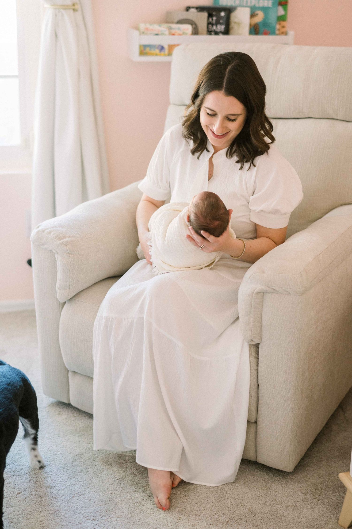 A woman sits in a gray armchair holding a baby while a dog watches nearby.