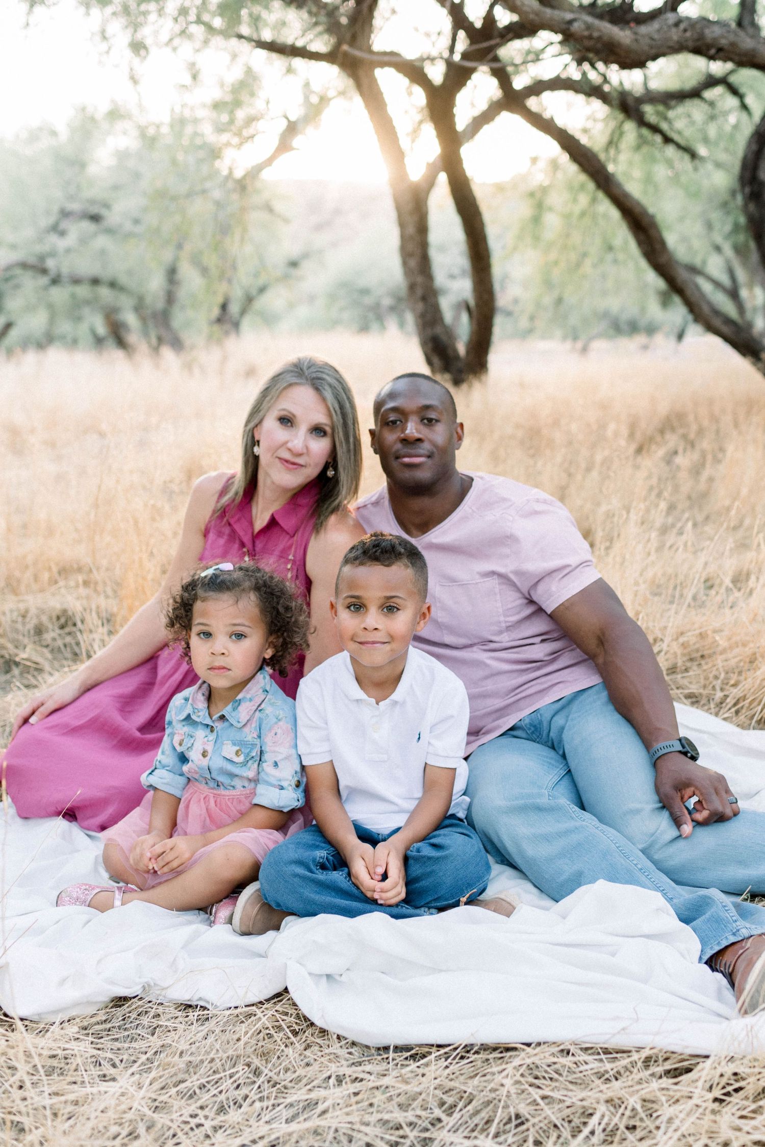 A family of four poses together on a white blanket in a sunlit field with trees.