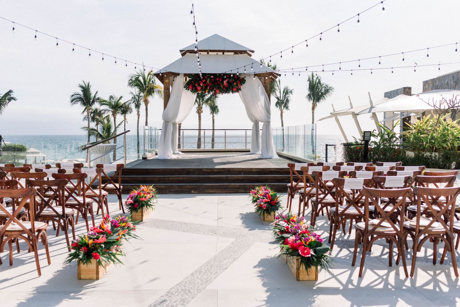 Beachfront wedding ceremony setup with white gazebo and wooden chairs under string lights.