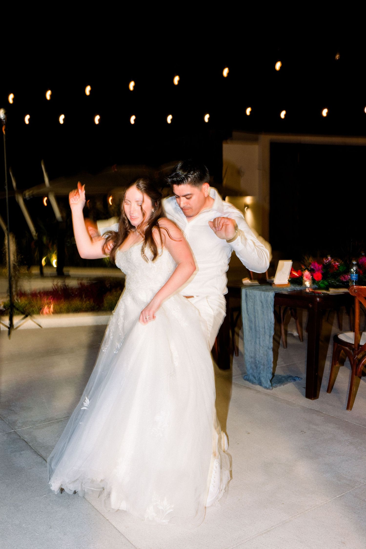 Newlyweds share a romantic dance moment under string lights at outdoor reception.