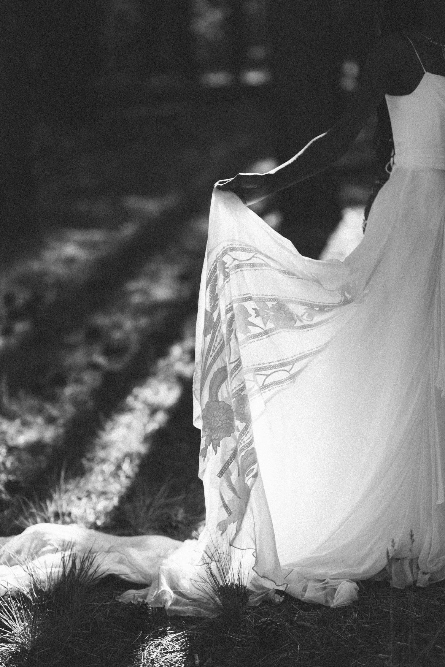 A black and white detail shot of a flowing white wedding dress hem against natural ground.