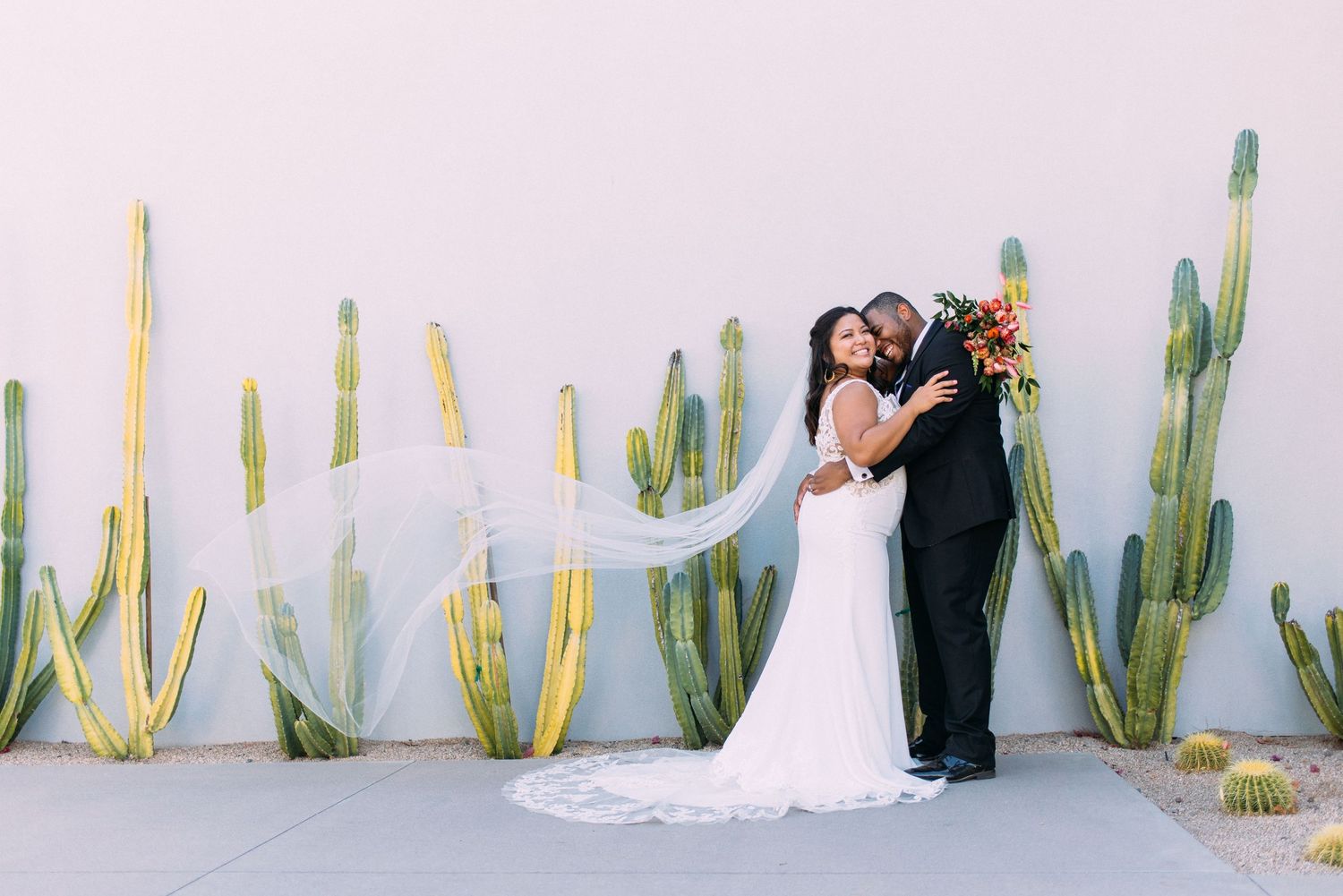 A couple in wedding attire poses together against a white wall with tall green cacti in the desert.
