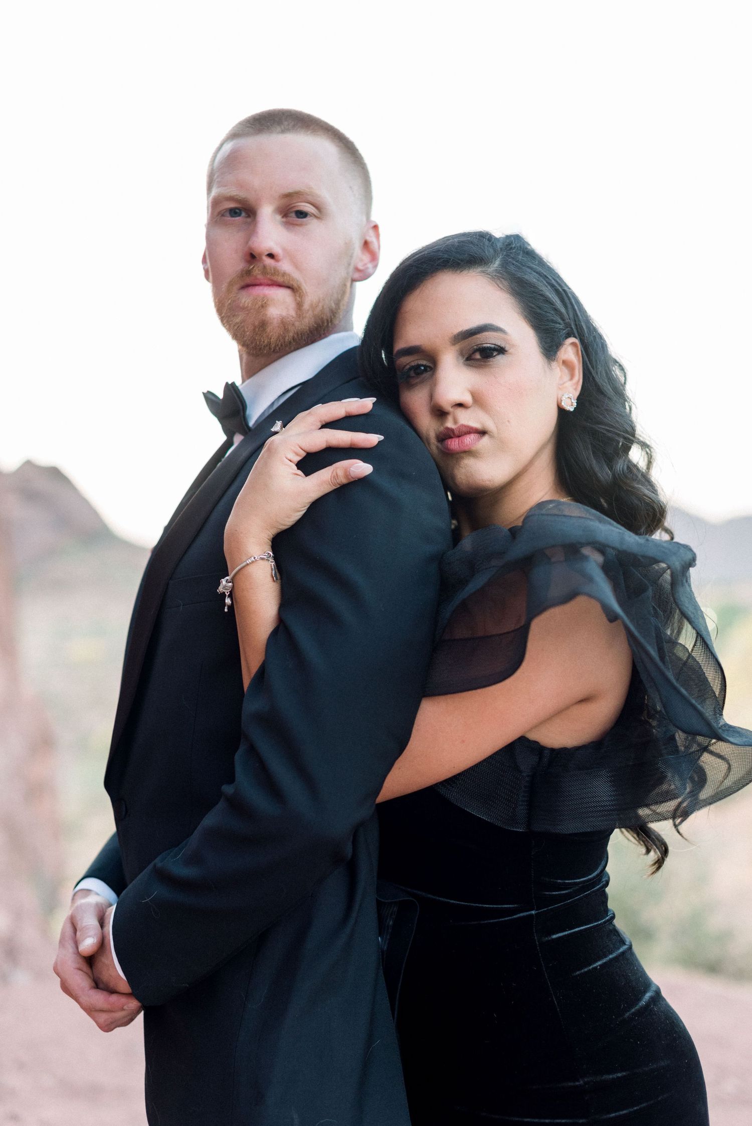 Stylish couple in formal wear poses against desert backdrop during sunset photoshoot.