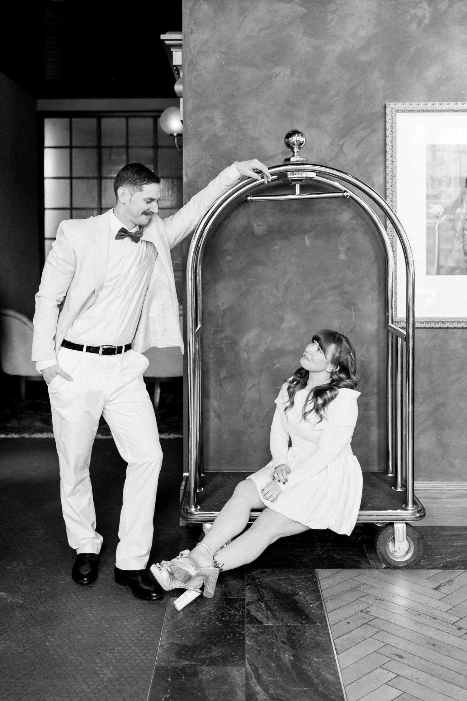 A black and white photo of a couple posing creatively with a vintage luggage cart.