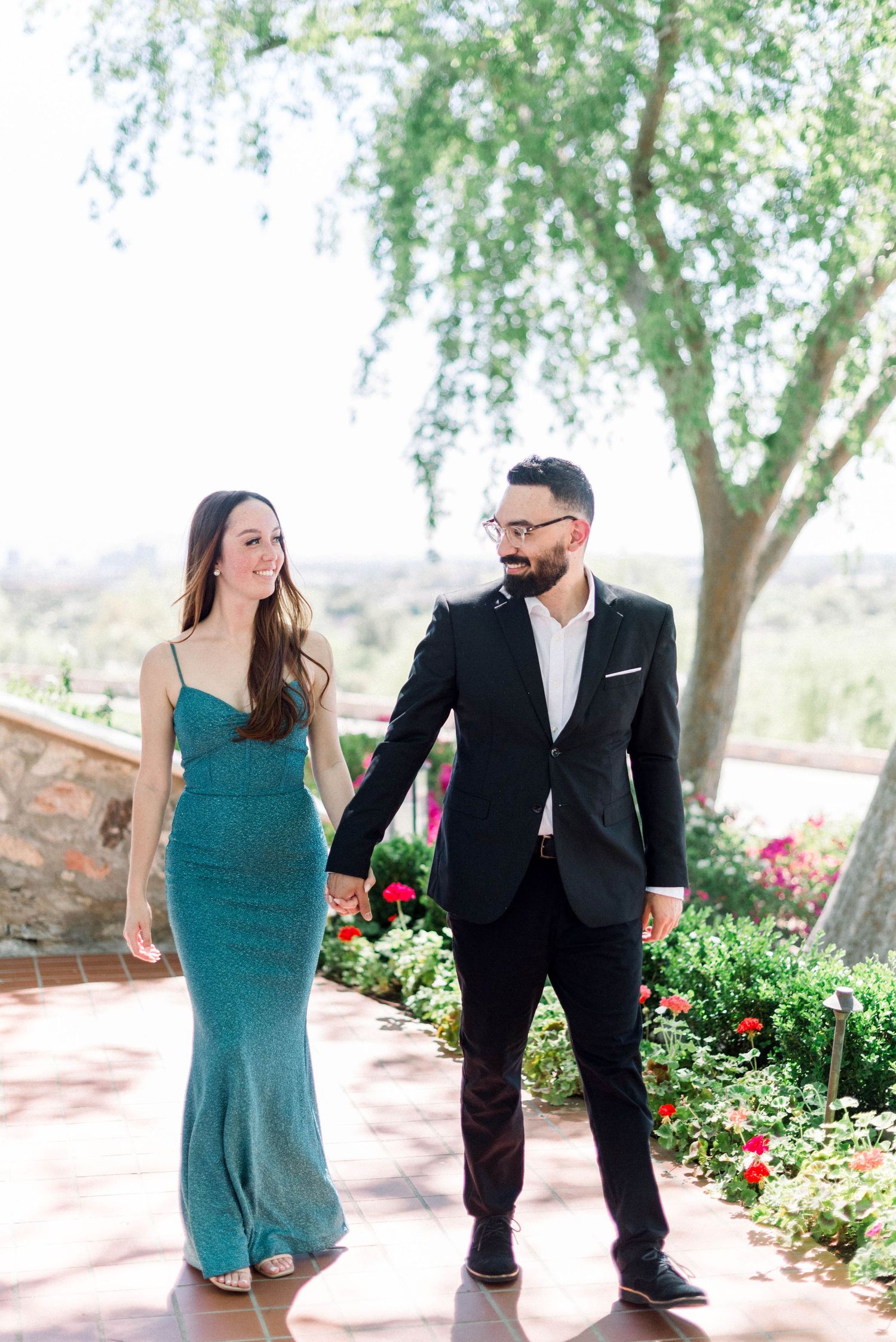 Couple strolls together on a sunny patio surrounded by flowering plants and trees.