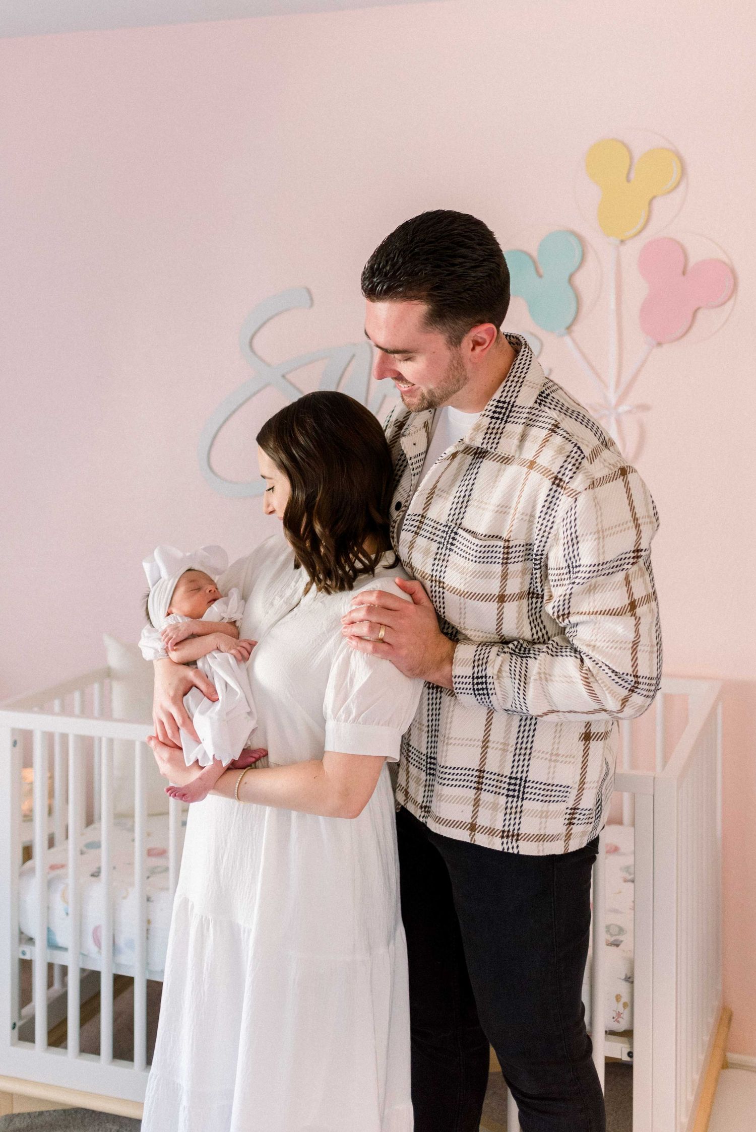 Parents hold their newborn baby in front of a pink nursery wall decorated with pastel heart balloons.