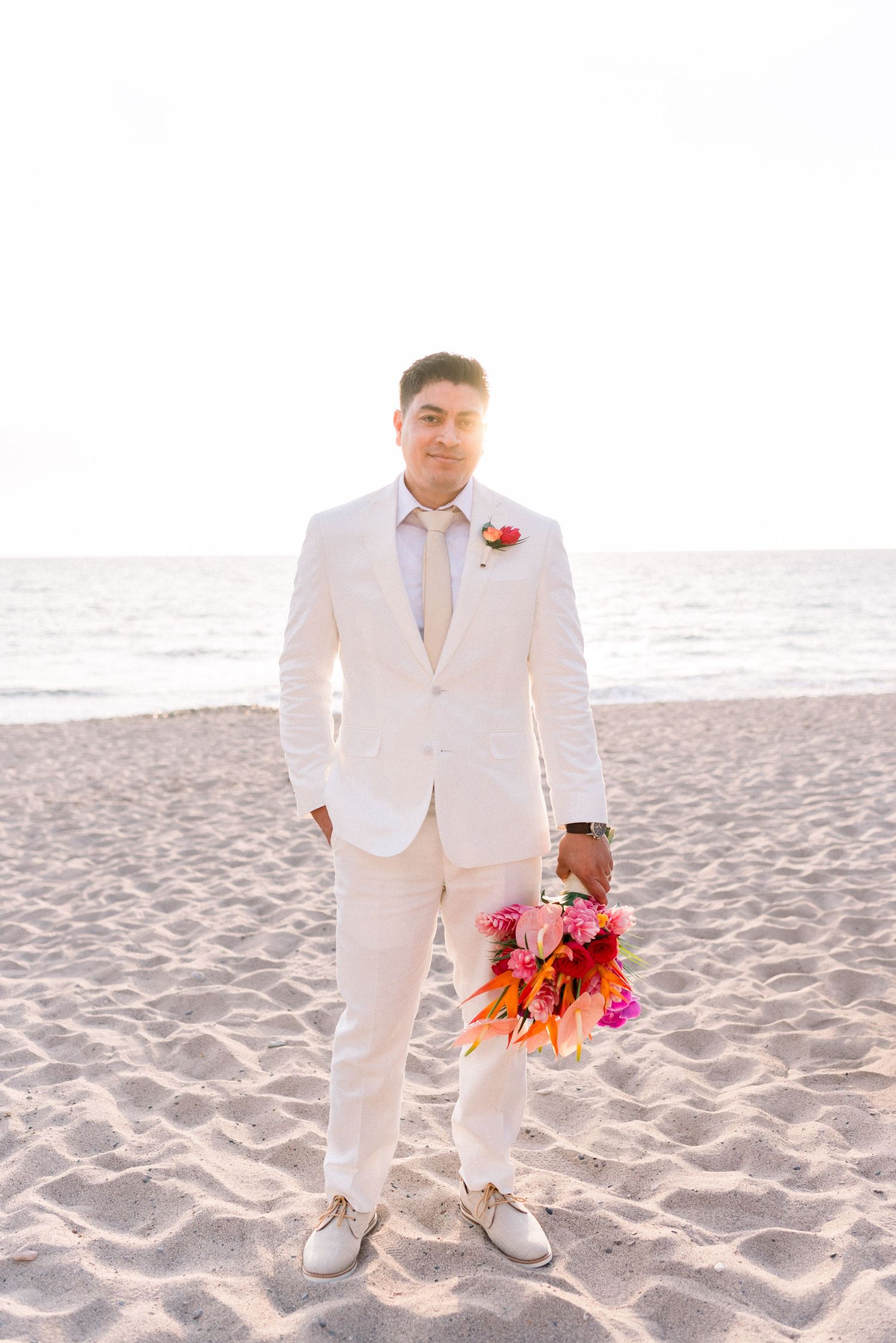 A person in a crisp white suit holds a vibrant tropical bouquet on a sunny beach.