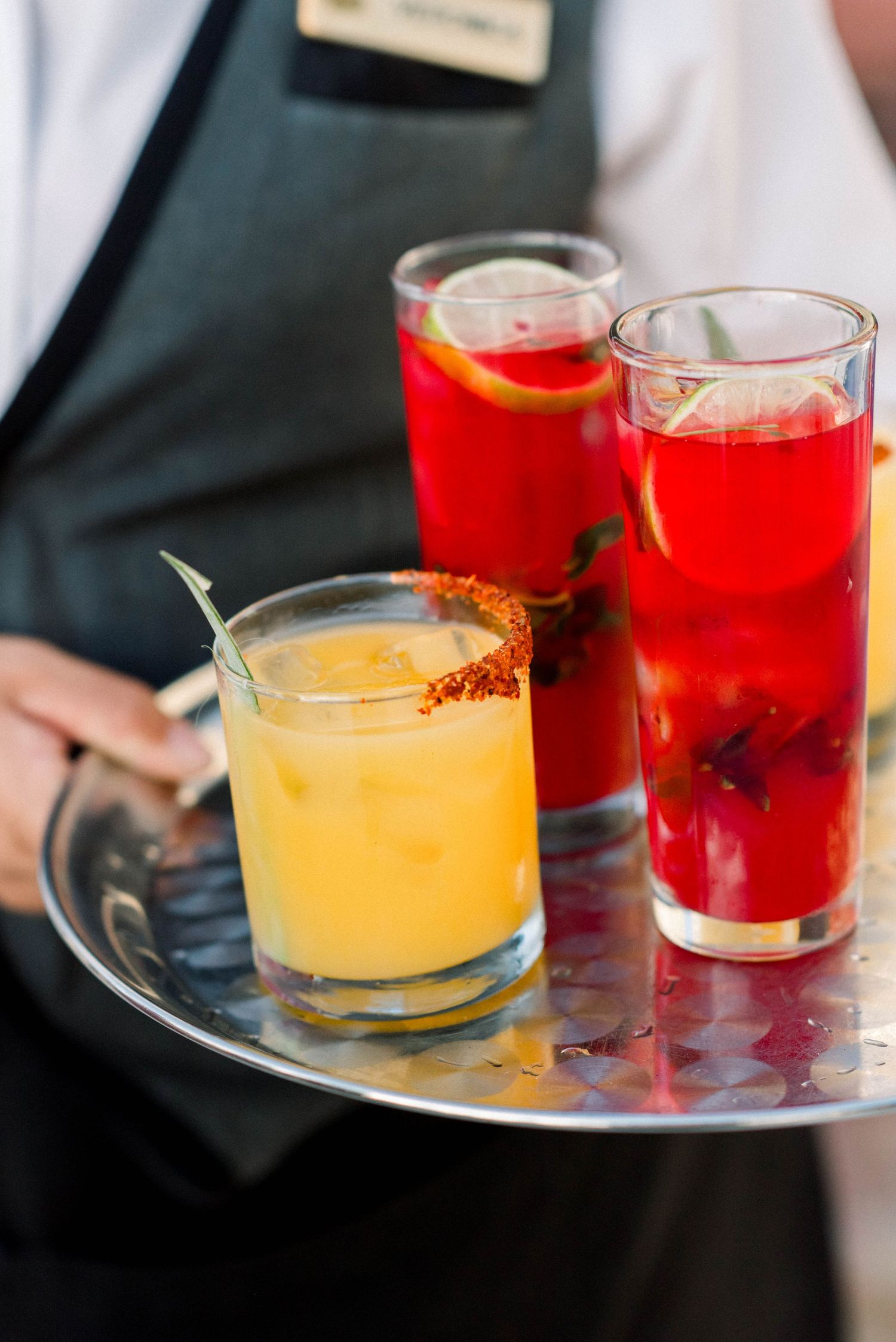 Colorful wedding cocktails served on a silver tray including yellow and red drinks with garnishes.