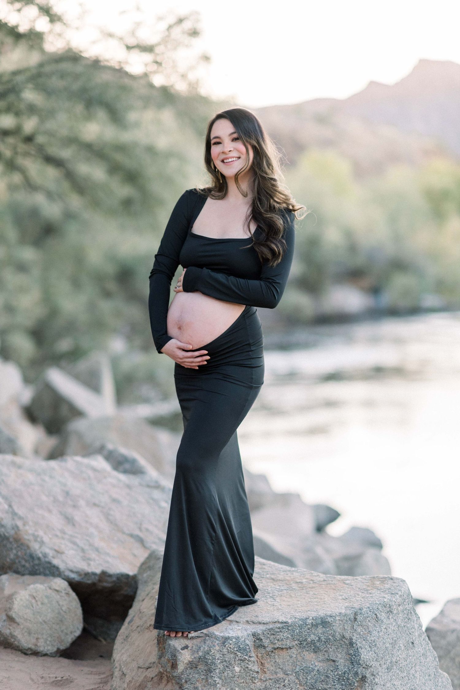 Joyful maternity portrait in black dress against rocky desert landscape.