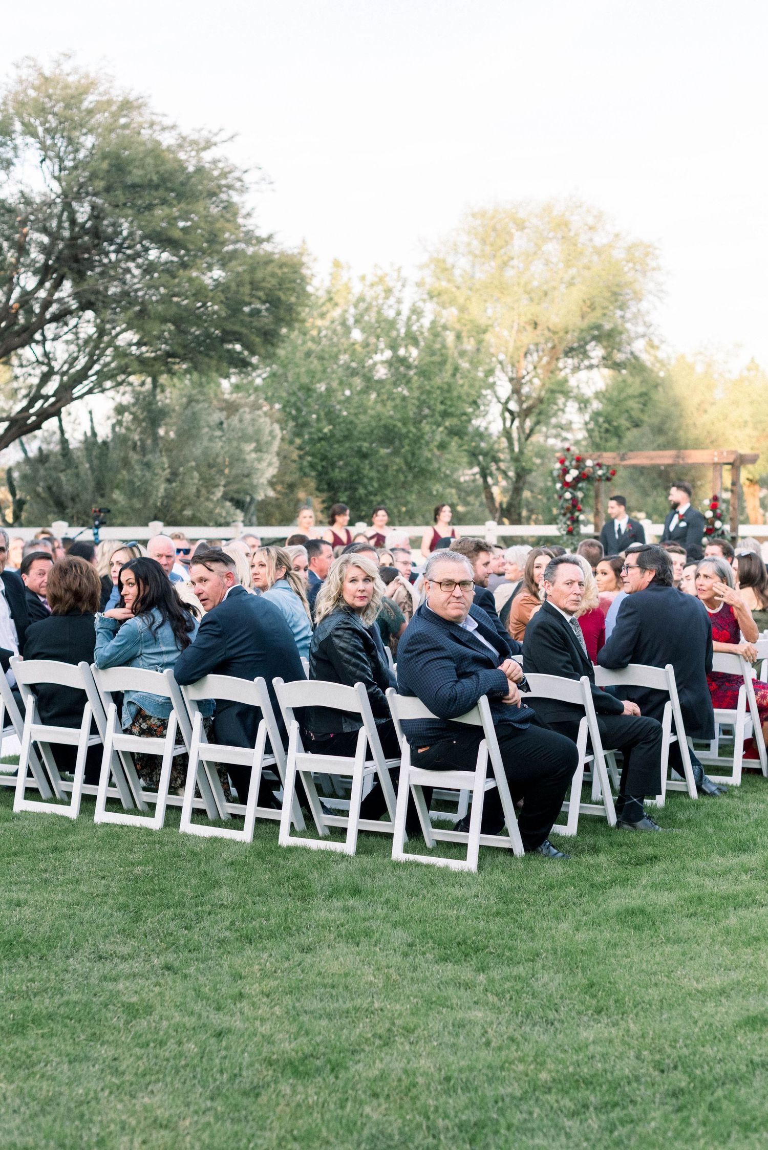 Outdoor wedding ceremony with guests seated in white folding chairs on a manicured lawn.
