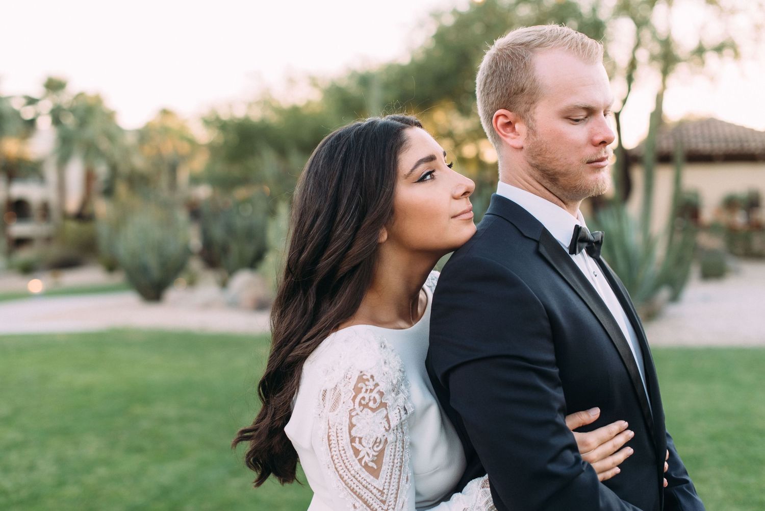 A romantic outdoor wedding portrait of a couple in formal attire against a desert landscape at sunset.