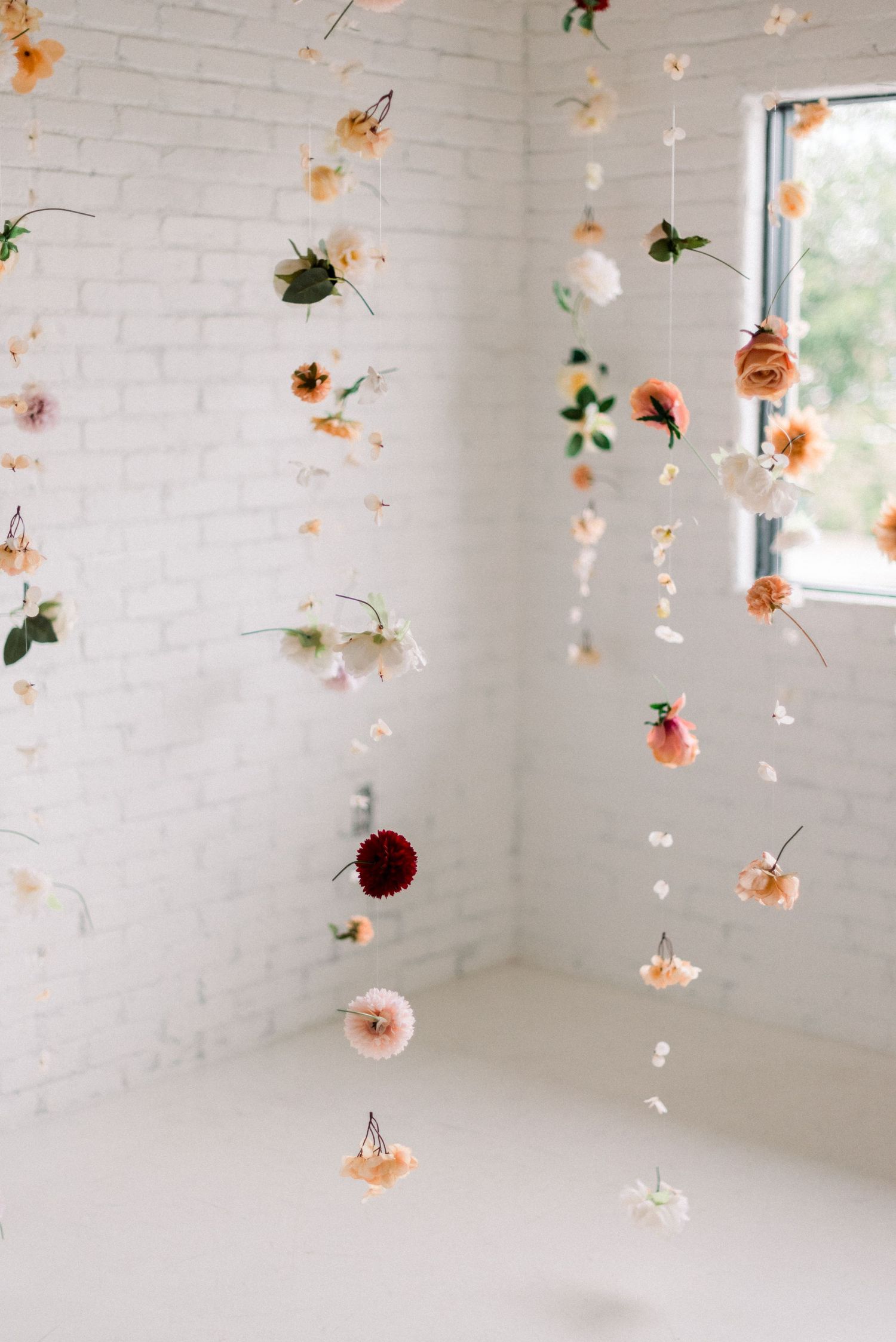 Whimsical hanging floral installation with roses and petals against white brick wall and window.