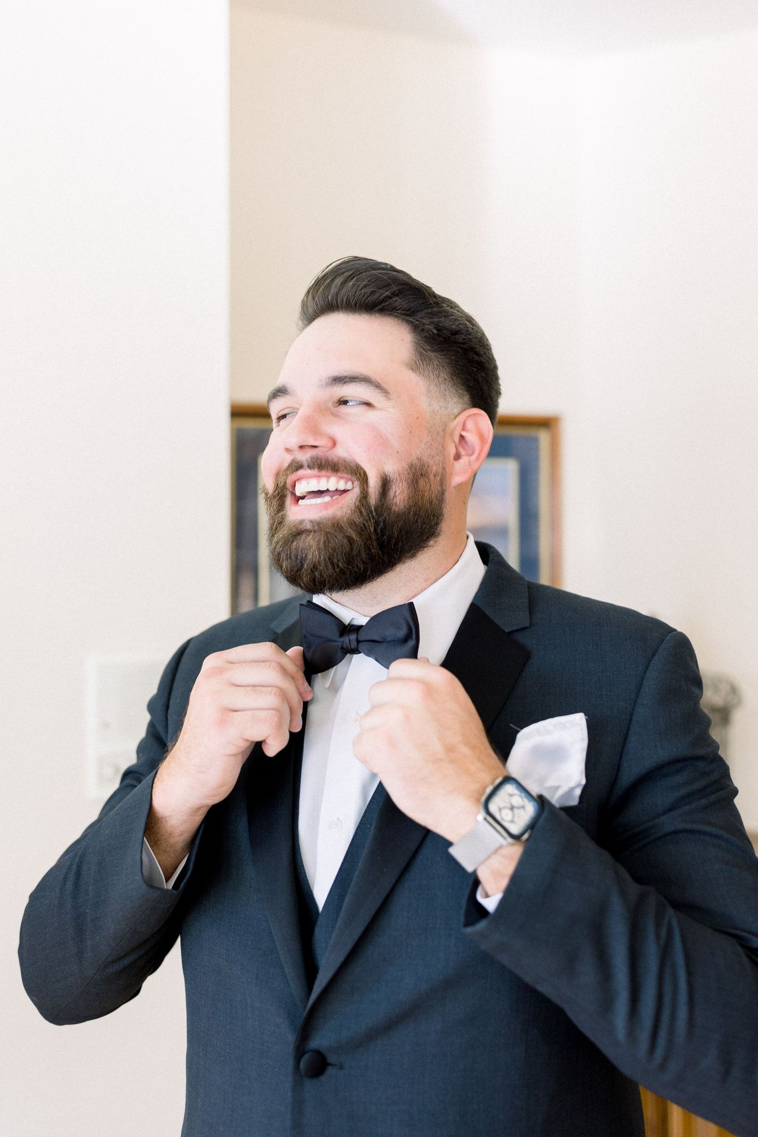 Smiling groom adjusts bowtie while wearing classic black tuxedo.