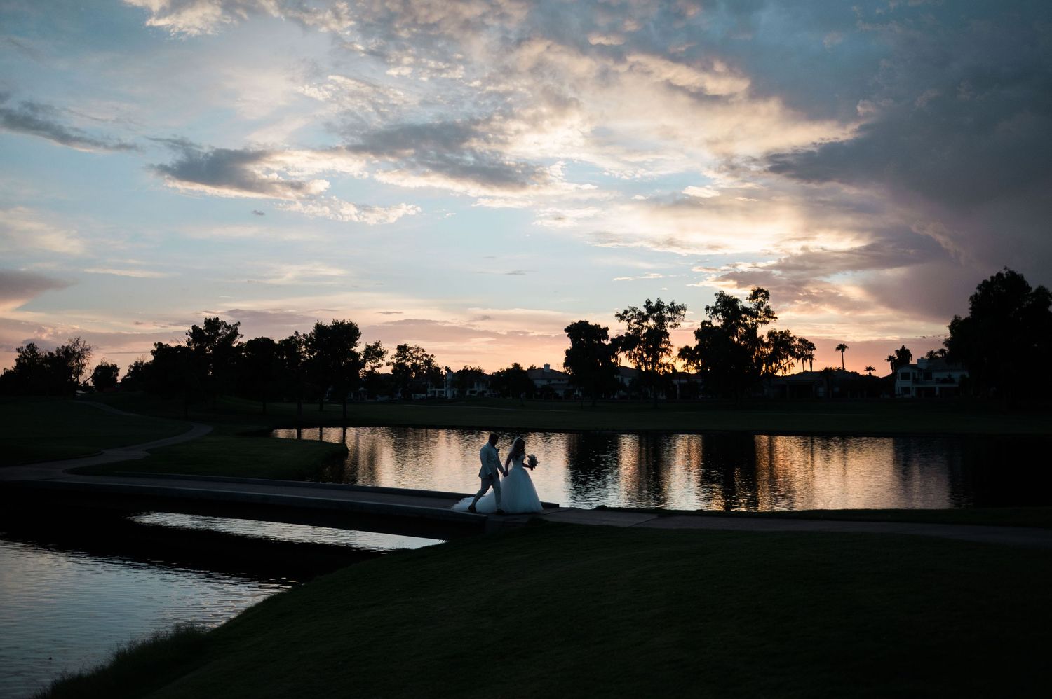 A tranquil lake reflects dramatic sunset clouds as silhouetted trees line the shoreline in a panoramic landscape view.