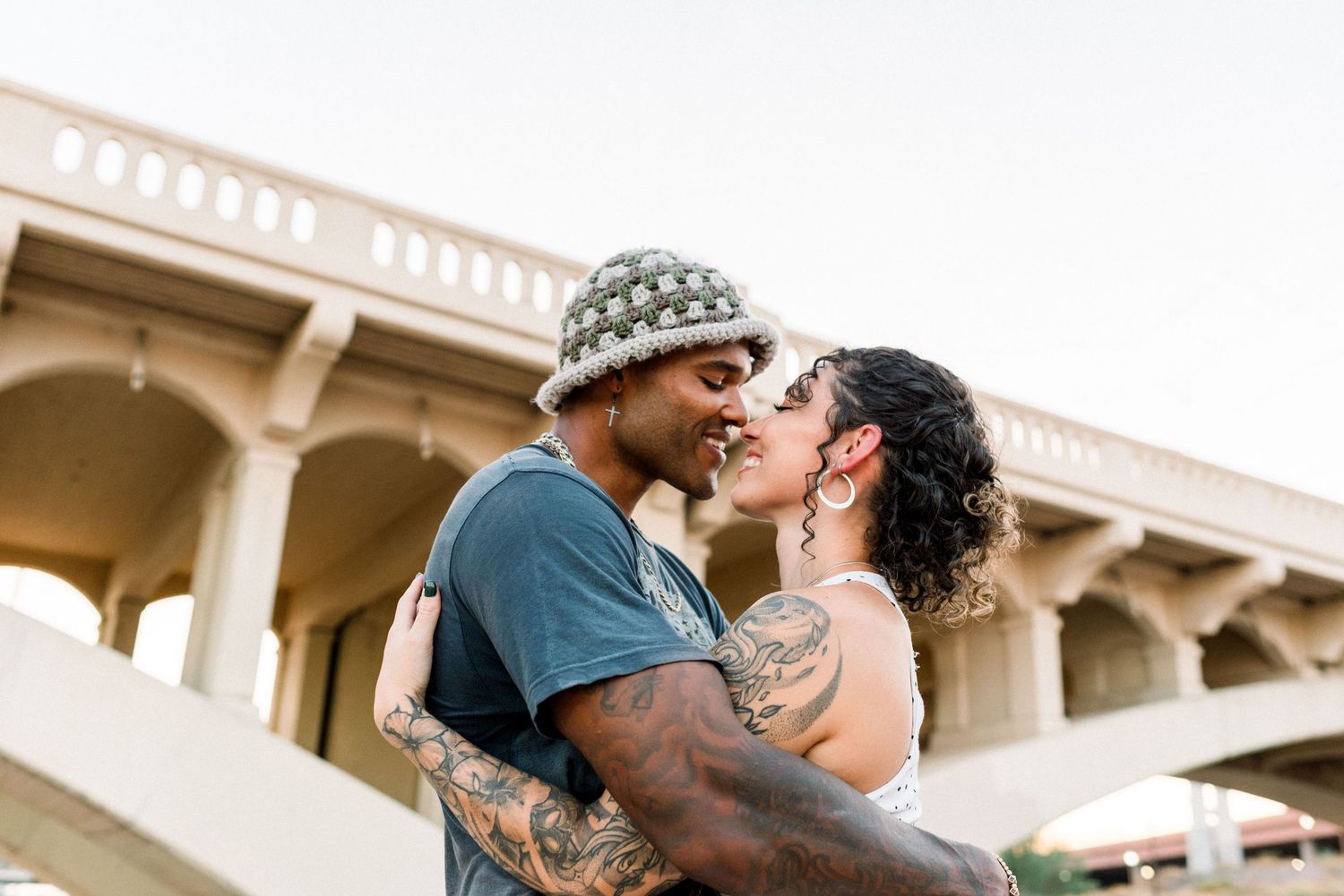 A couple shares a romantic kiss outdoors in a sun-drenched setting with architectural details in the background.