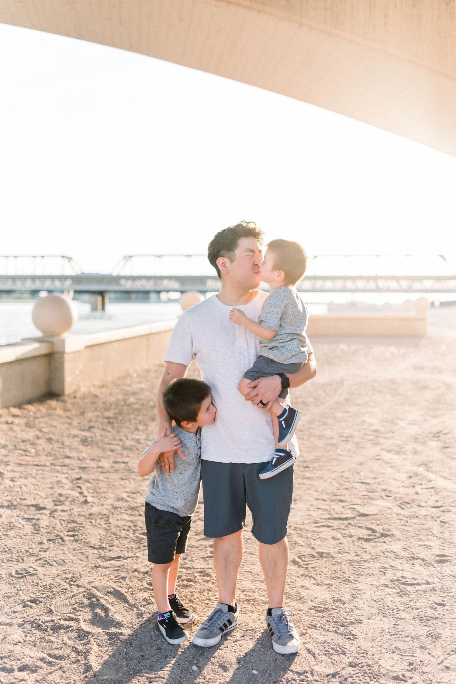 A parent and two children share a tender moment on a sandy beach at sunset with a bridge in the background.