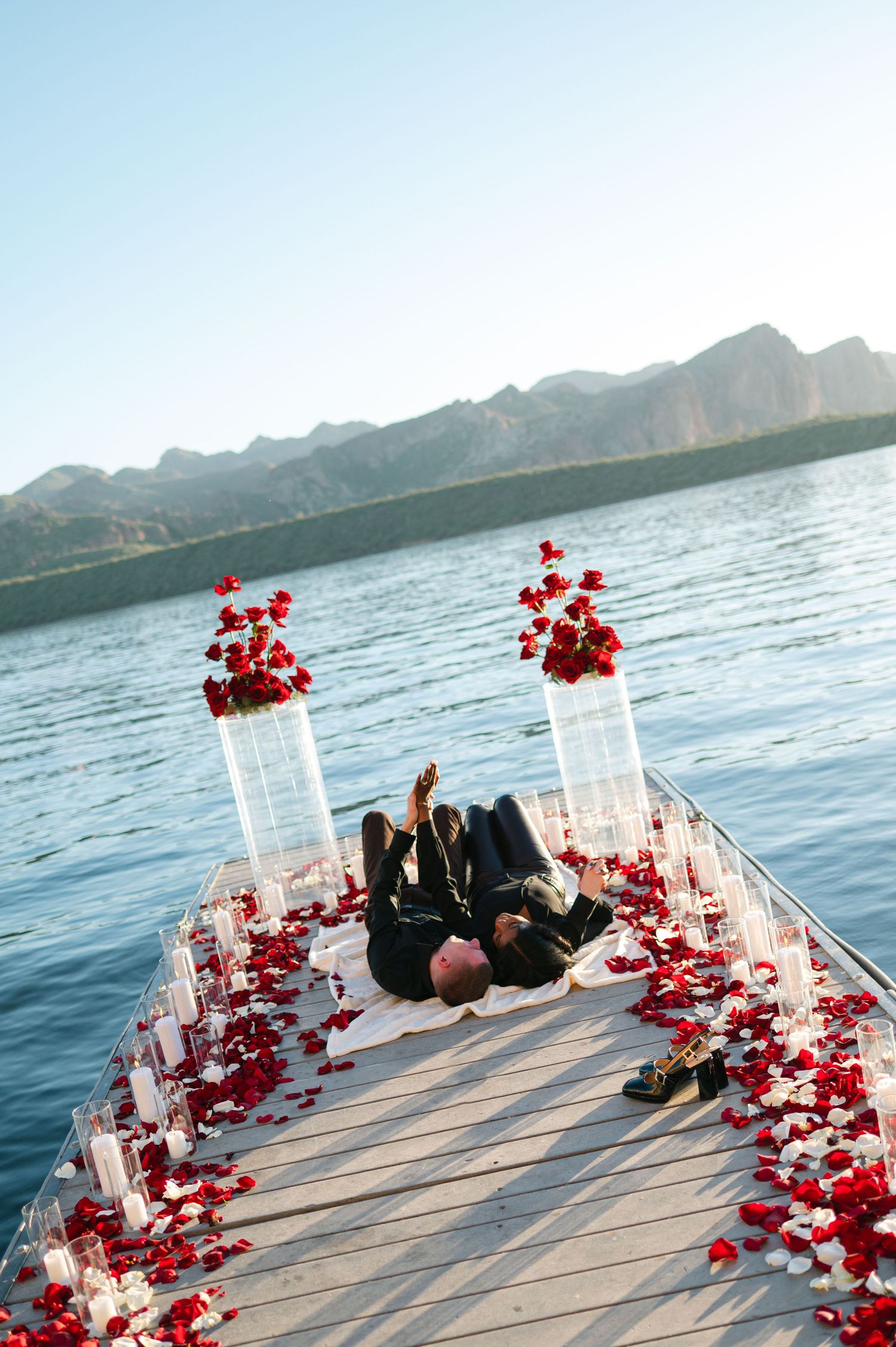 Romantic proposal setup on a lakeside dock decorated with red rose petals and white lanterns at sunset.